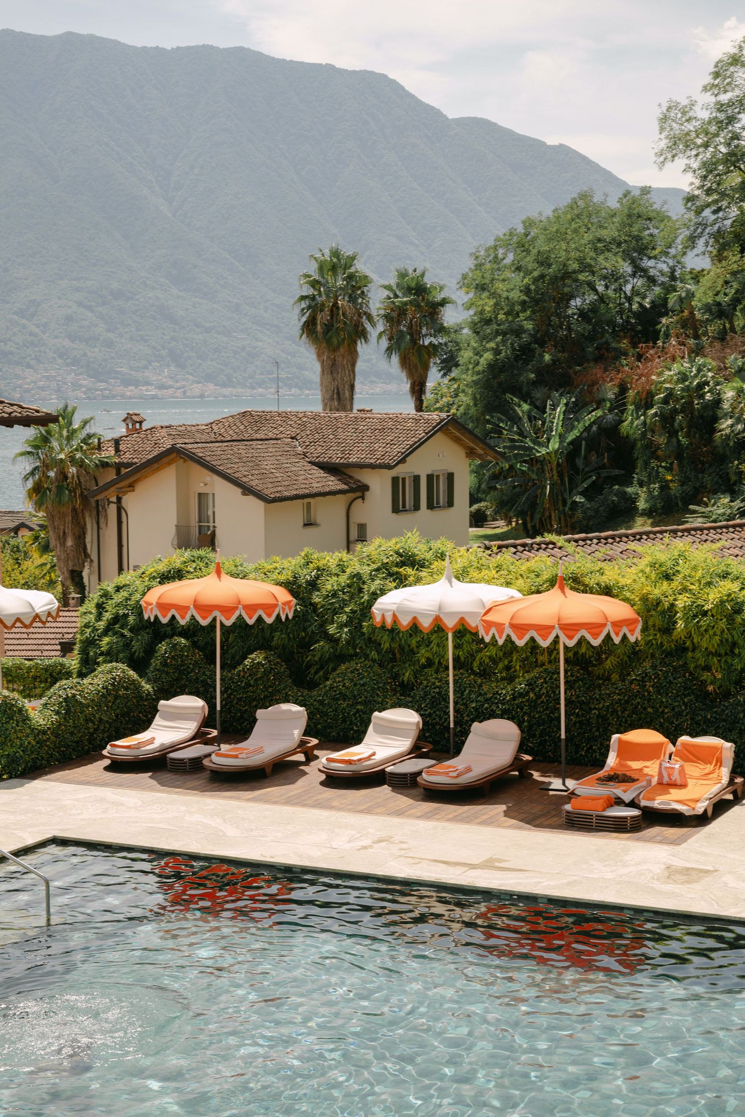 Luxury pool with orange-fringed umbrellas and lounge chairs overlooking a villa and mountains at an Italian lakeside resort.