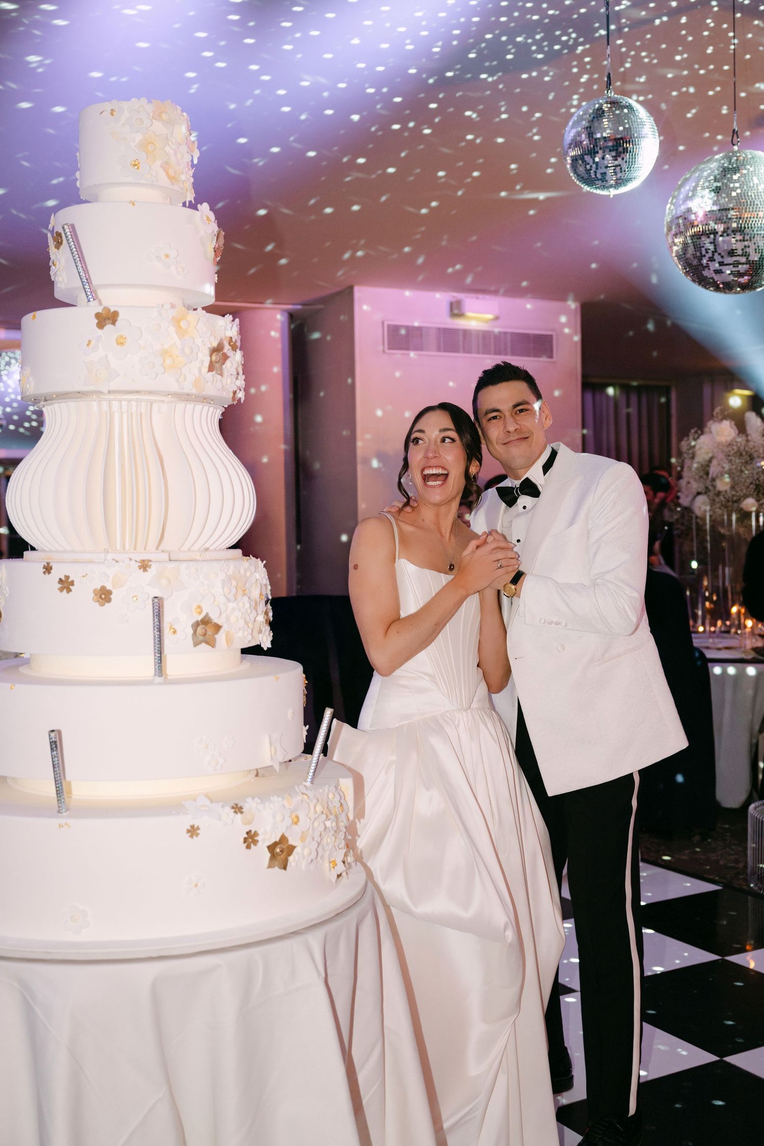 Joyful bride and groom posing beside tall white star-decorated wedding cake with confetti falling.