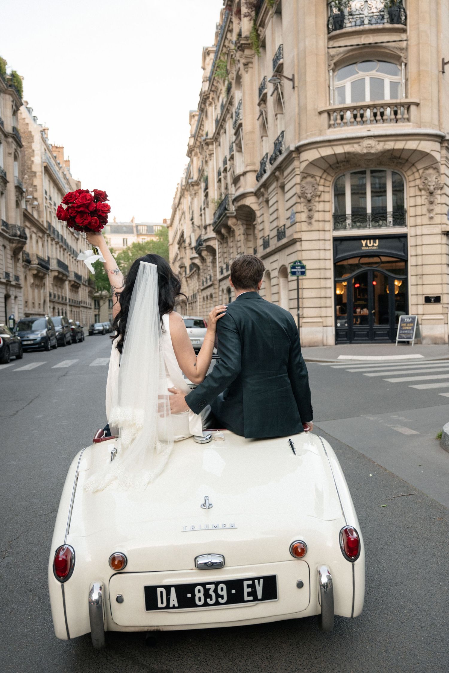 Newlyweds riding in a vintage white Triumph convertible through Paris streets, bride holding red rose bouquet.