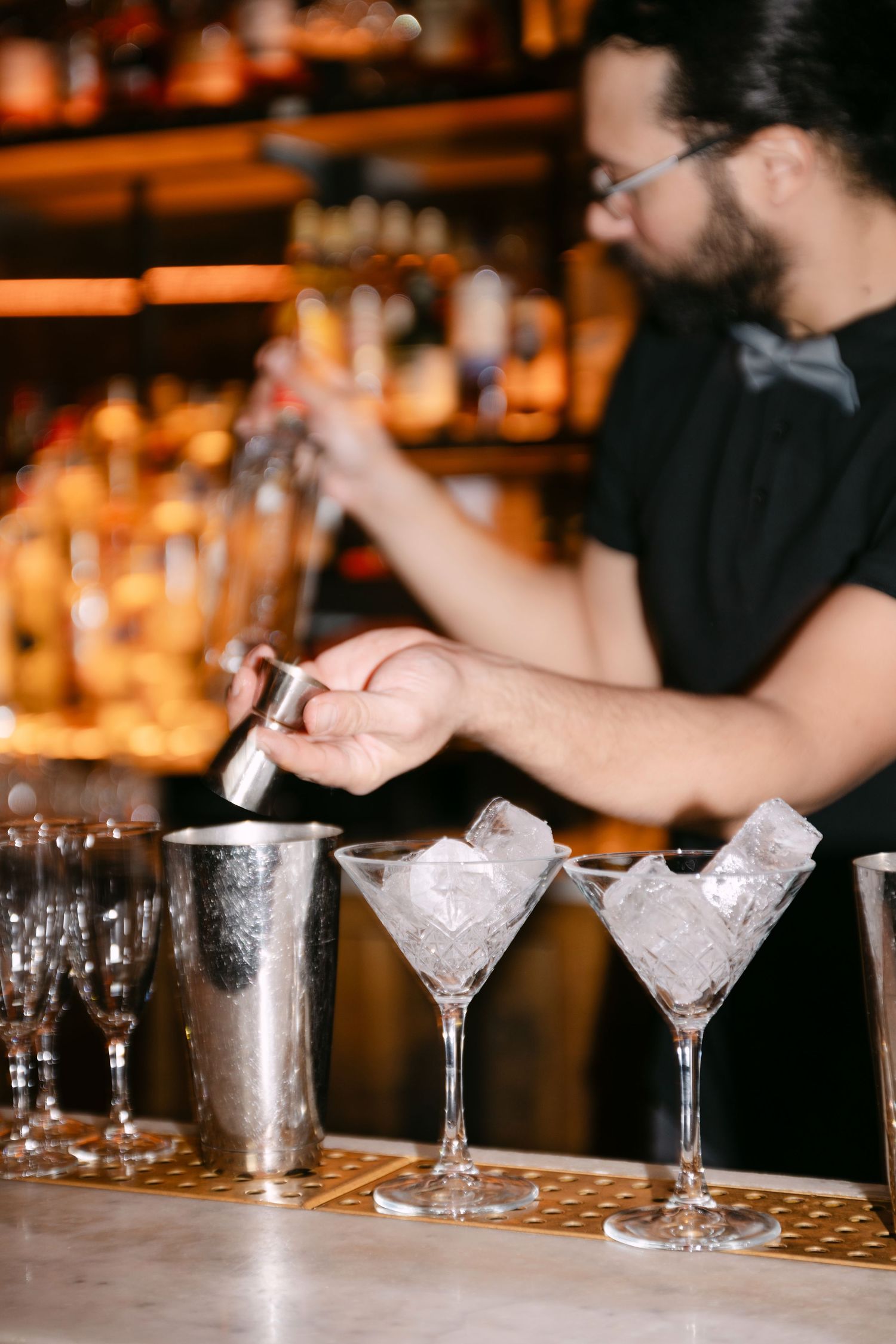 Bearded bartender in bow tie preparing cocktails with ice-filled martini glasses.