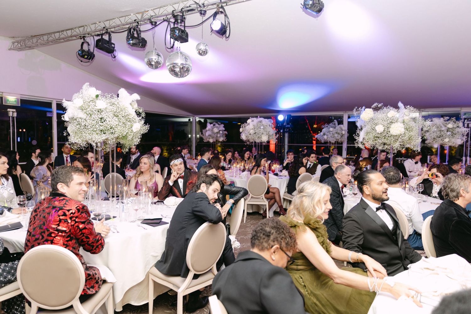 Elegantly dressed wedding guests seated at round tables beneath tall floral centerpieces in a tented reception venue.