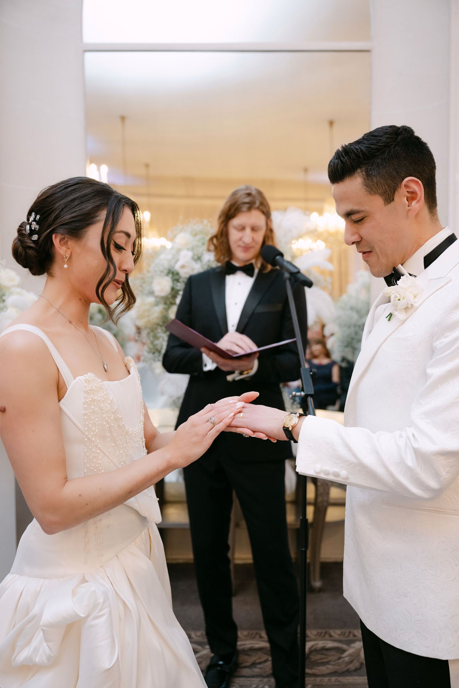 Groom placing wedding ring on brides finger as officiant reads from book during indoor ceremony.