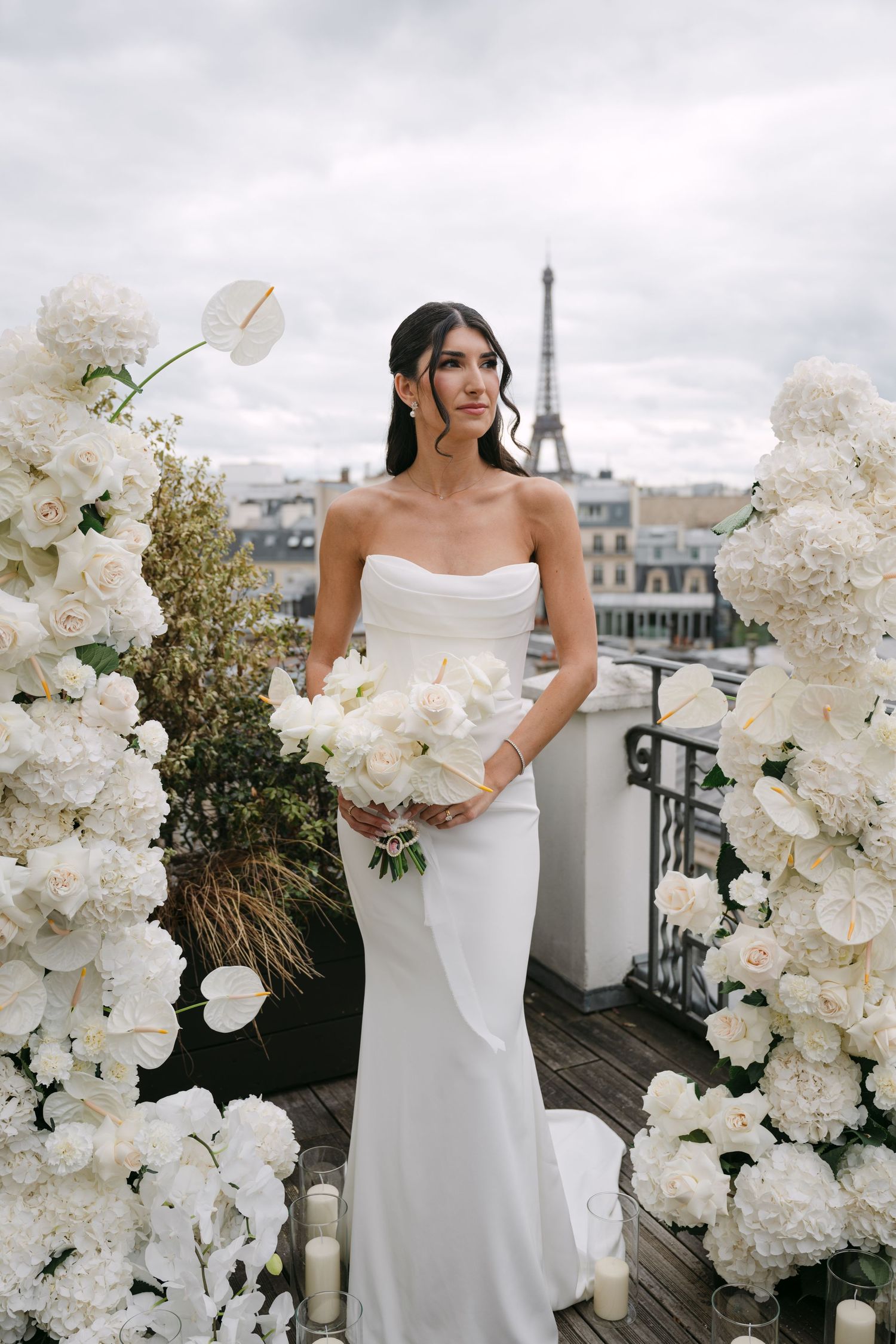 Bride holding white bouquet stands between floral arrangements on Parisian rooftop with Eiffel Tower view.