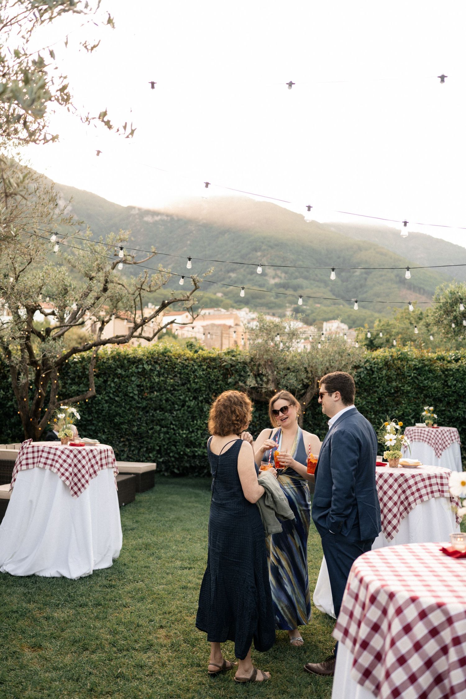 Guests mingle at an outdoor Italian wedding reception with checkered tablecloths and string lights against mountain backdrop.