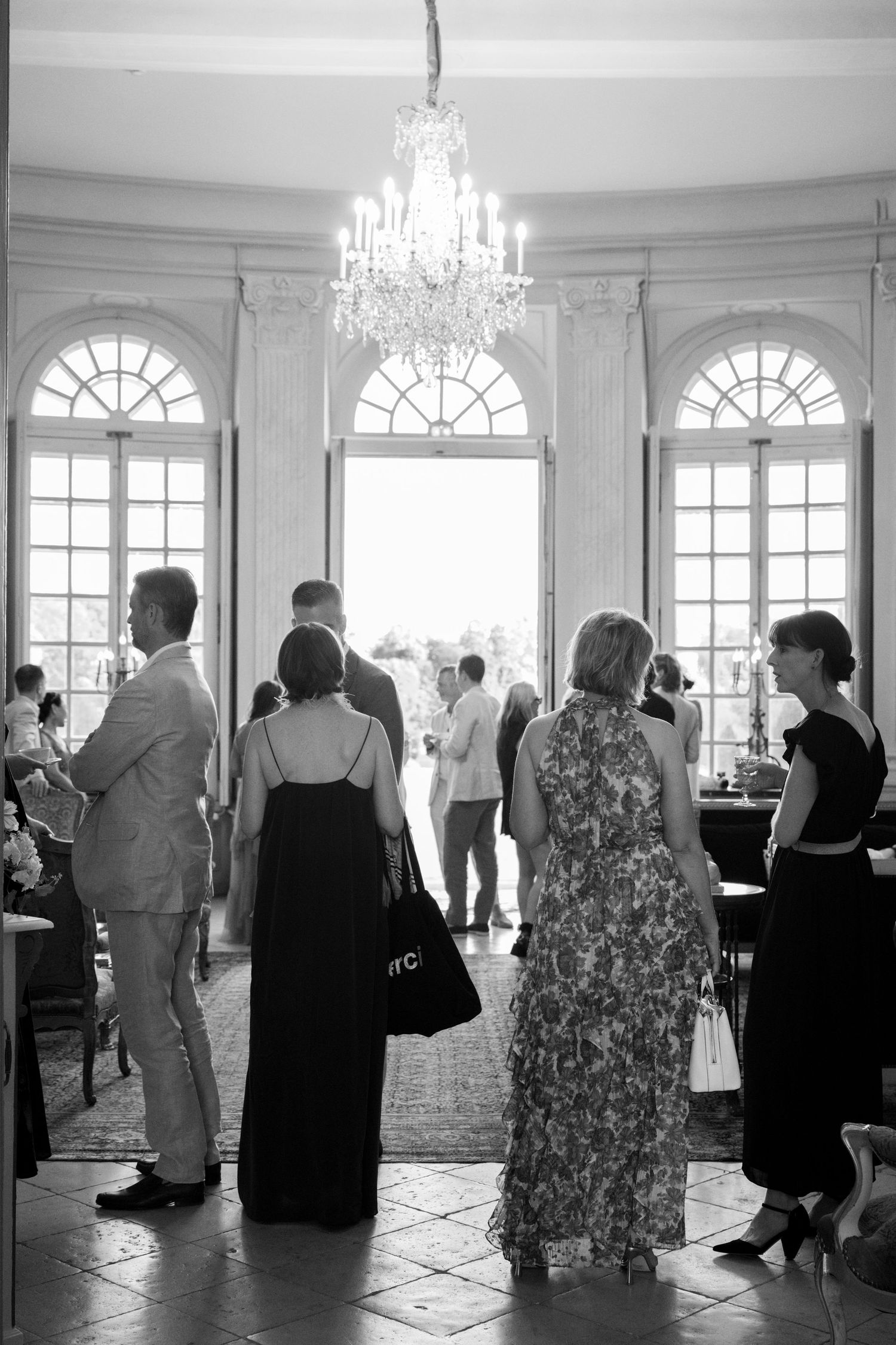 Black and white photo of elegantly dressed wedding guests gathered under a crystal chandelier in a grand French salon.