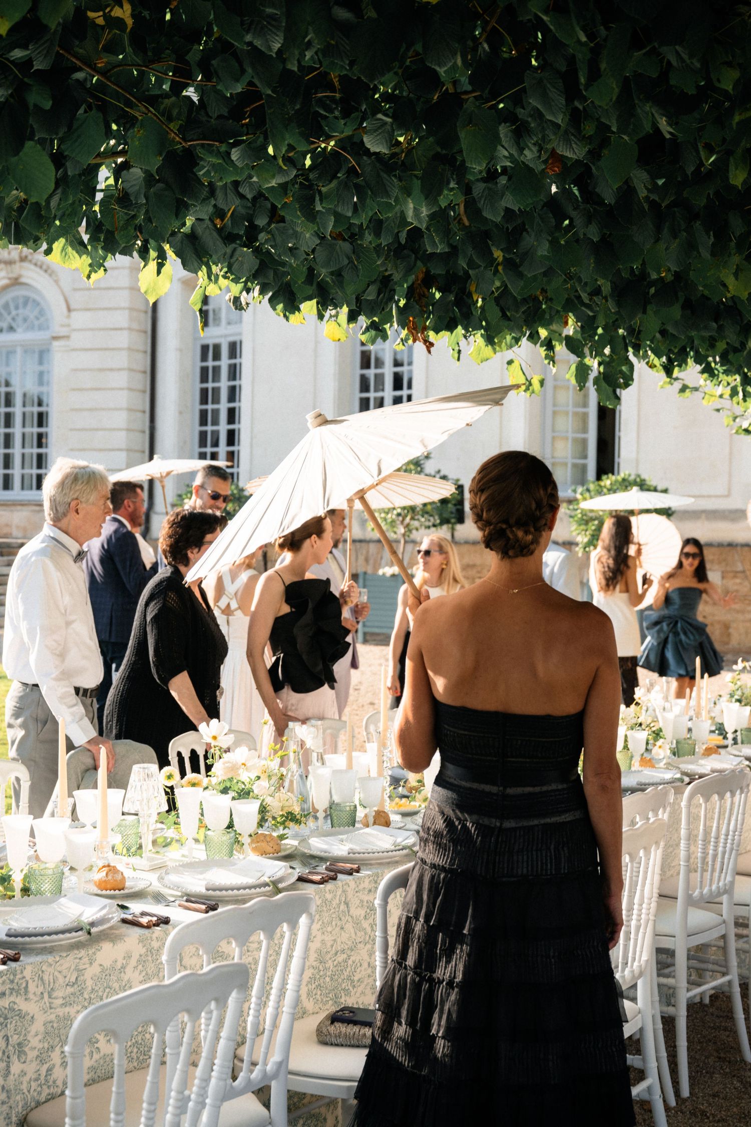 Wedding guests mingling with parasols at an outdoor garden reception beside a grand French estate.