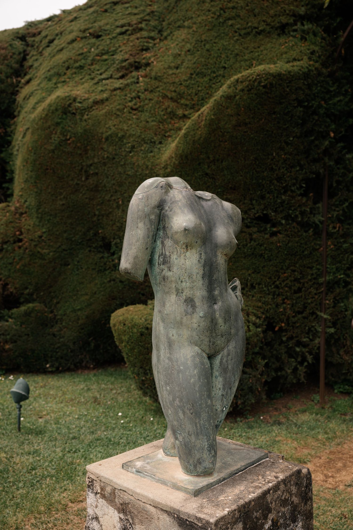 Headless bronze female torso sculpture on stone pedestal against manicured green hedge garden.