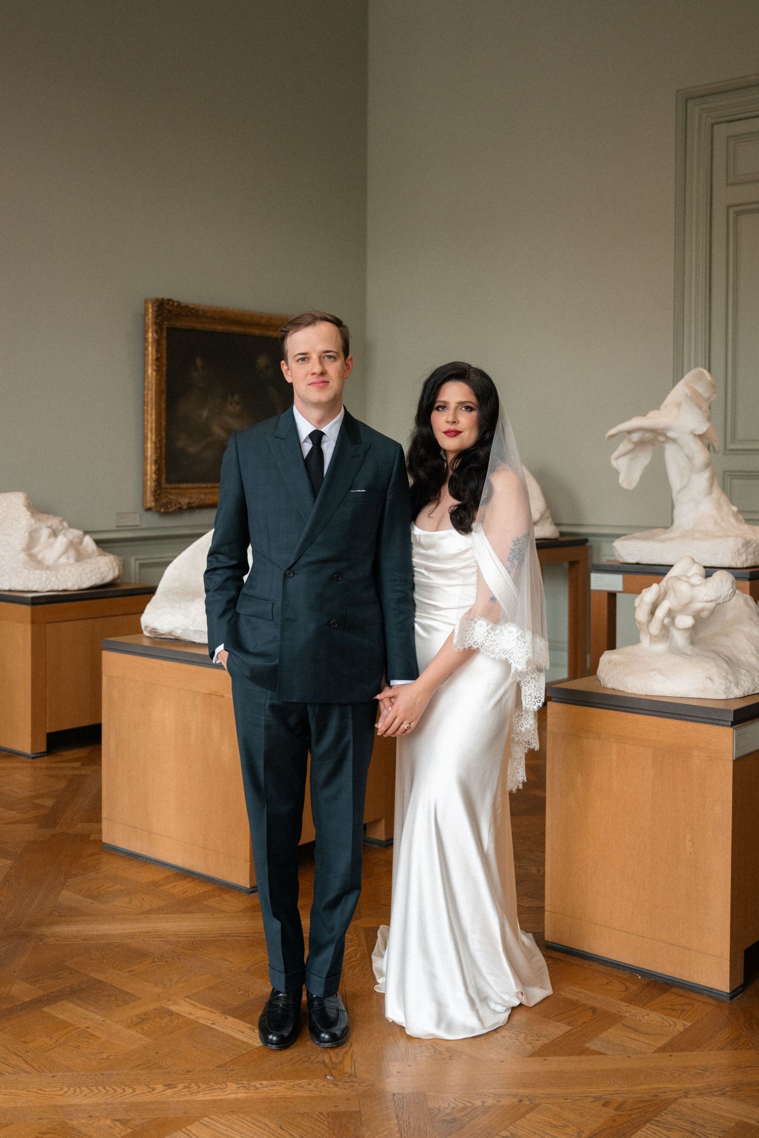 Bride and groom pose formally among classical sculptures in a grand museum gallery, wearing a white satin gown and dark suit.