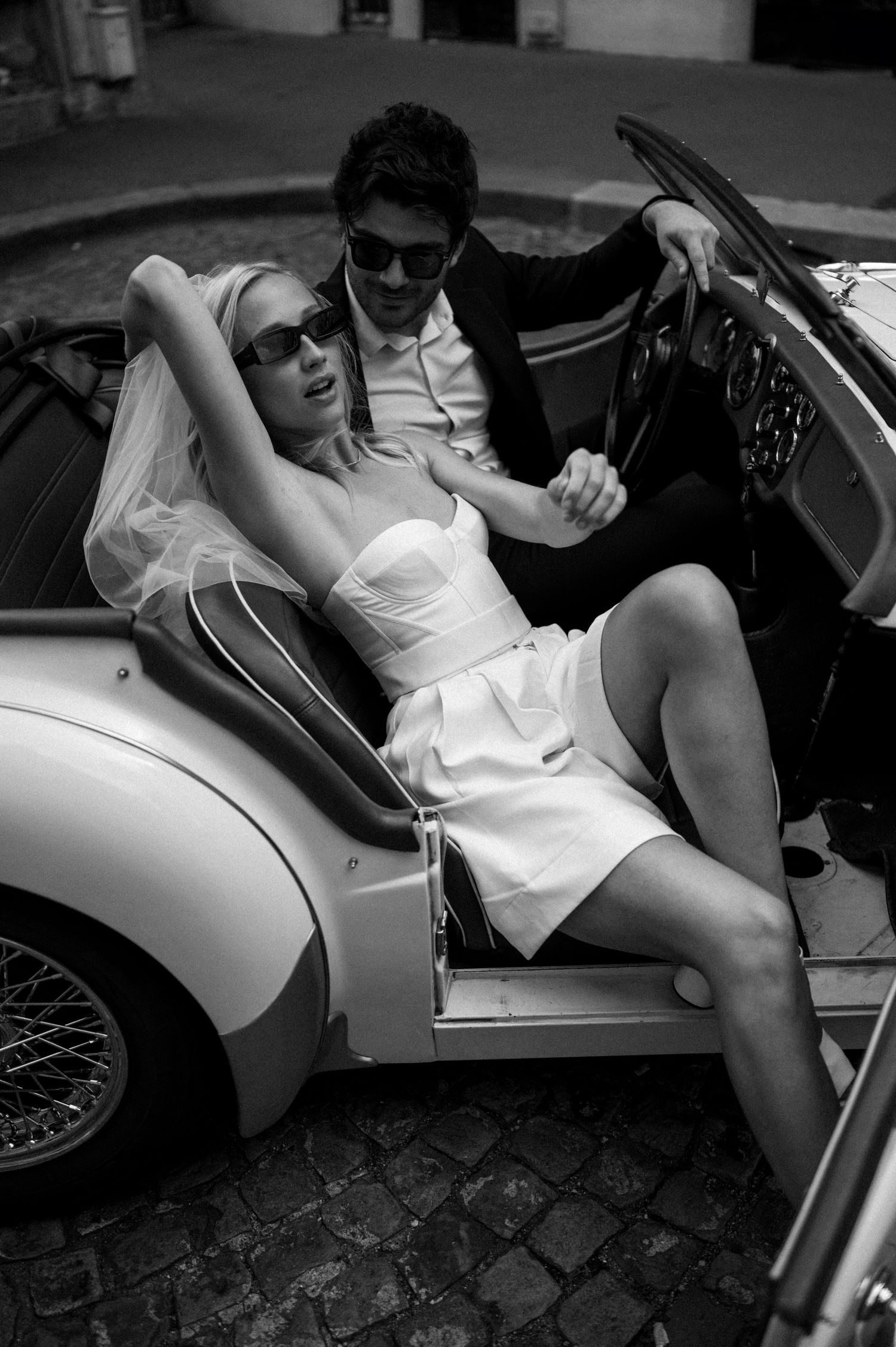 Black and white photo of bride in short dress and veil posing with groom in vintage convertible.