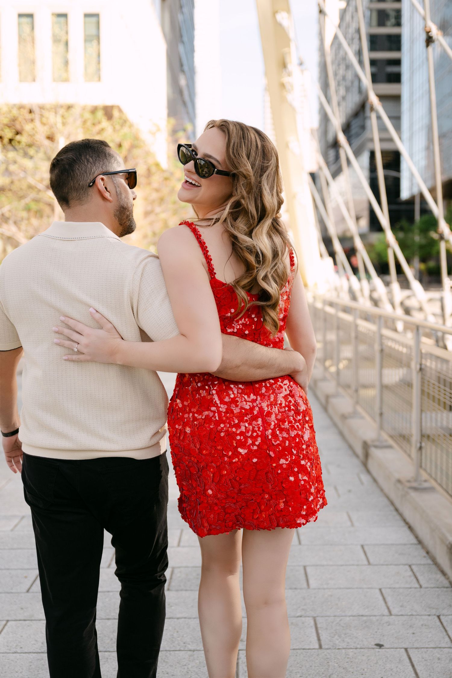 Stylish couple in sunglasses walk arm in arm on city bridge, woman wearing red sequin mini dress.