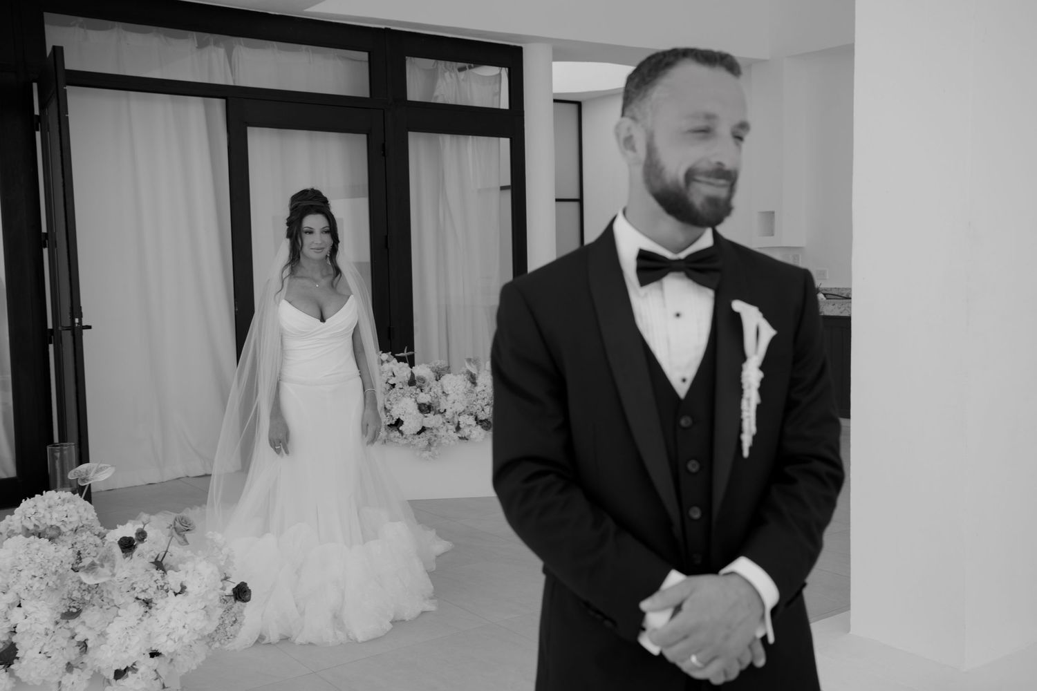 Black and white photo of a groom in tuxedo experiencing first look moment as bride in wedding gown approaches from behind.