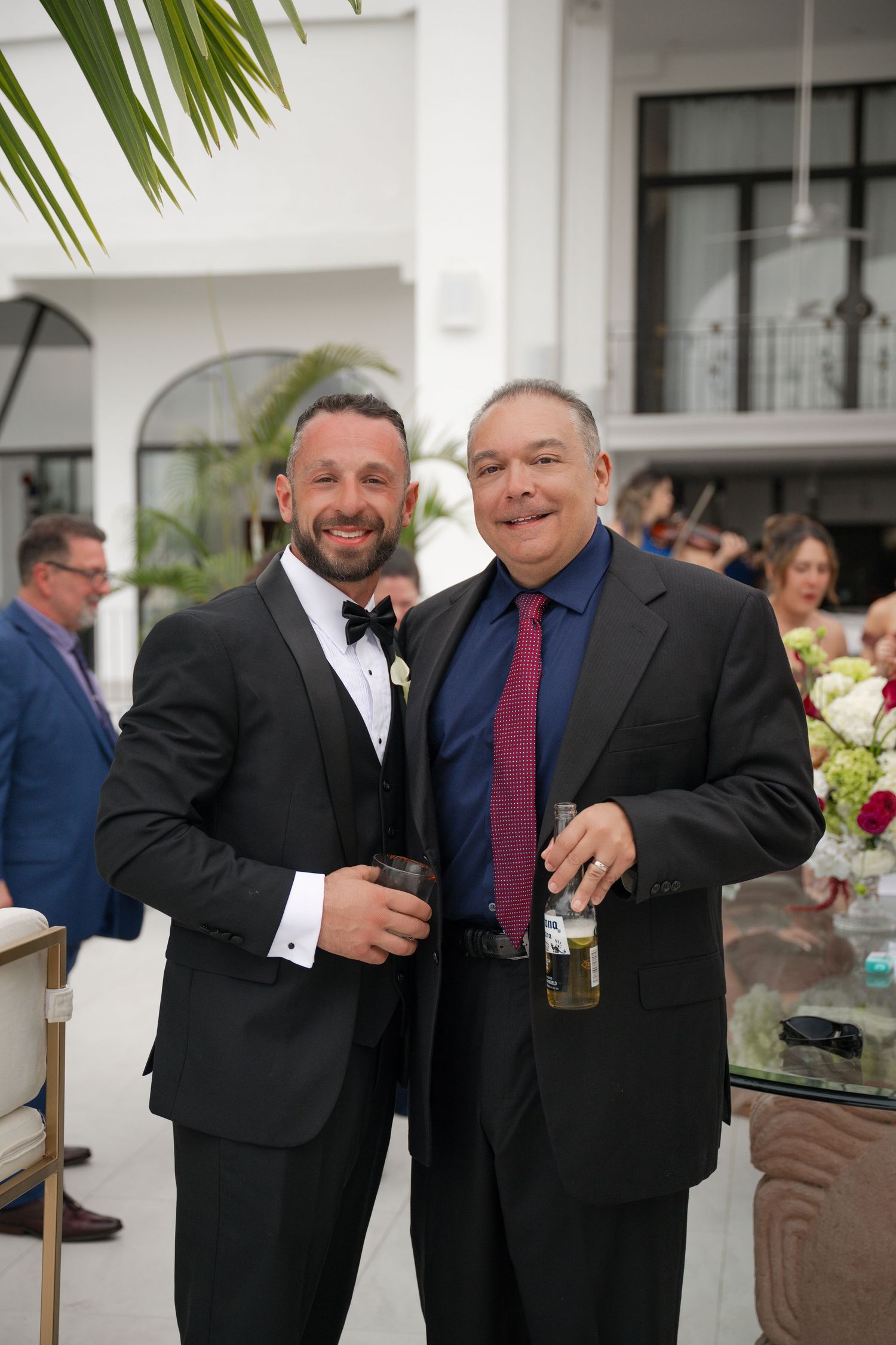 Two men in suits smile and hold drinks at an outdoor wedding reception.