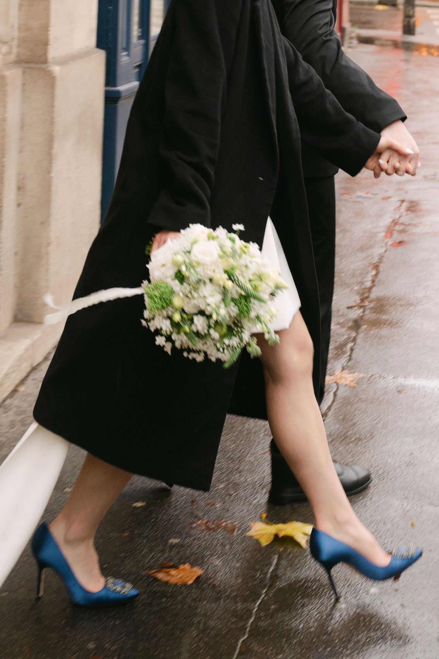 Bride in black coat and blue heels walks on wet pavement holding a white floral bouquet.