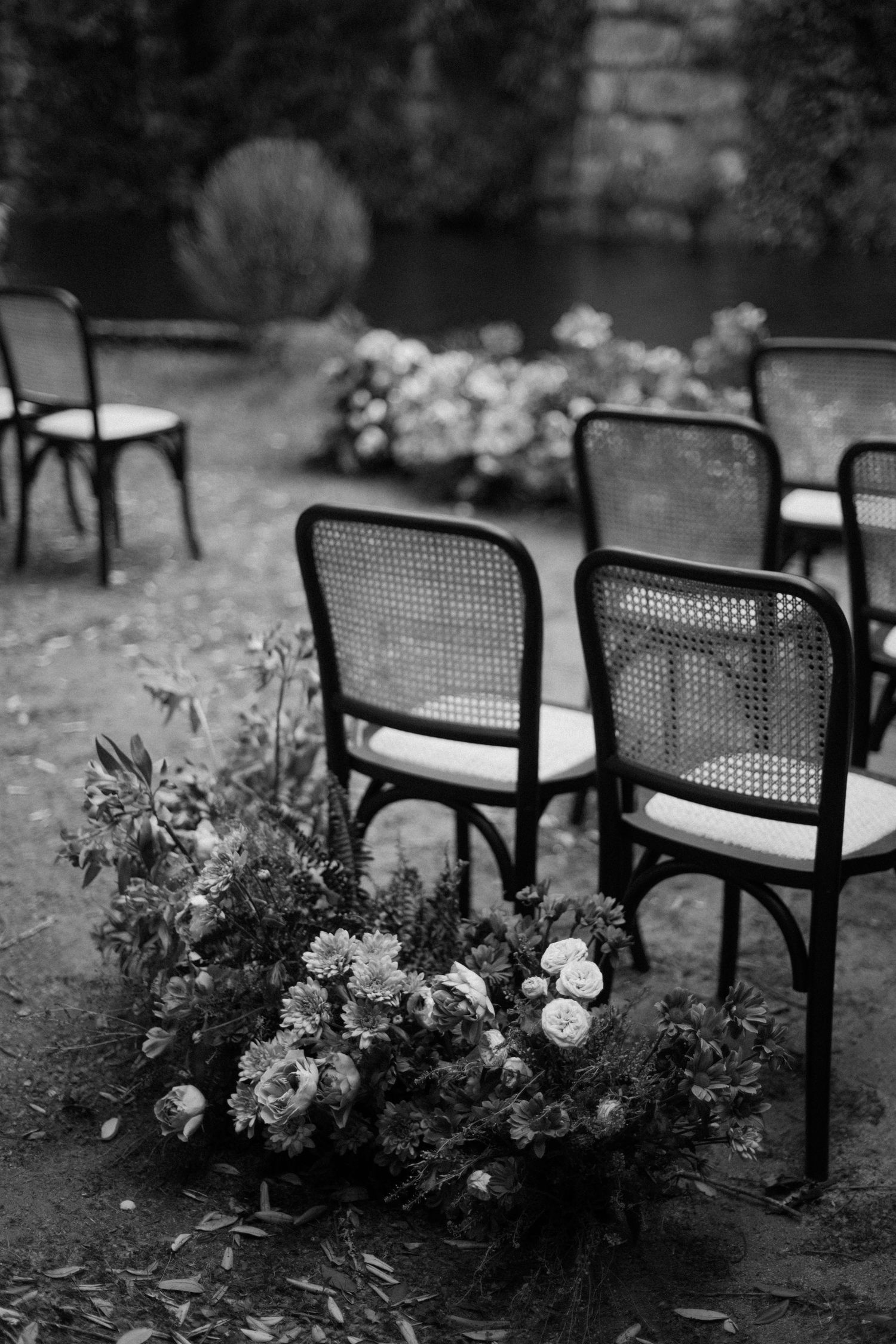 Black and white photo of elegant cane chairs and floral arrangement at an outdoor wedding ceremony.