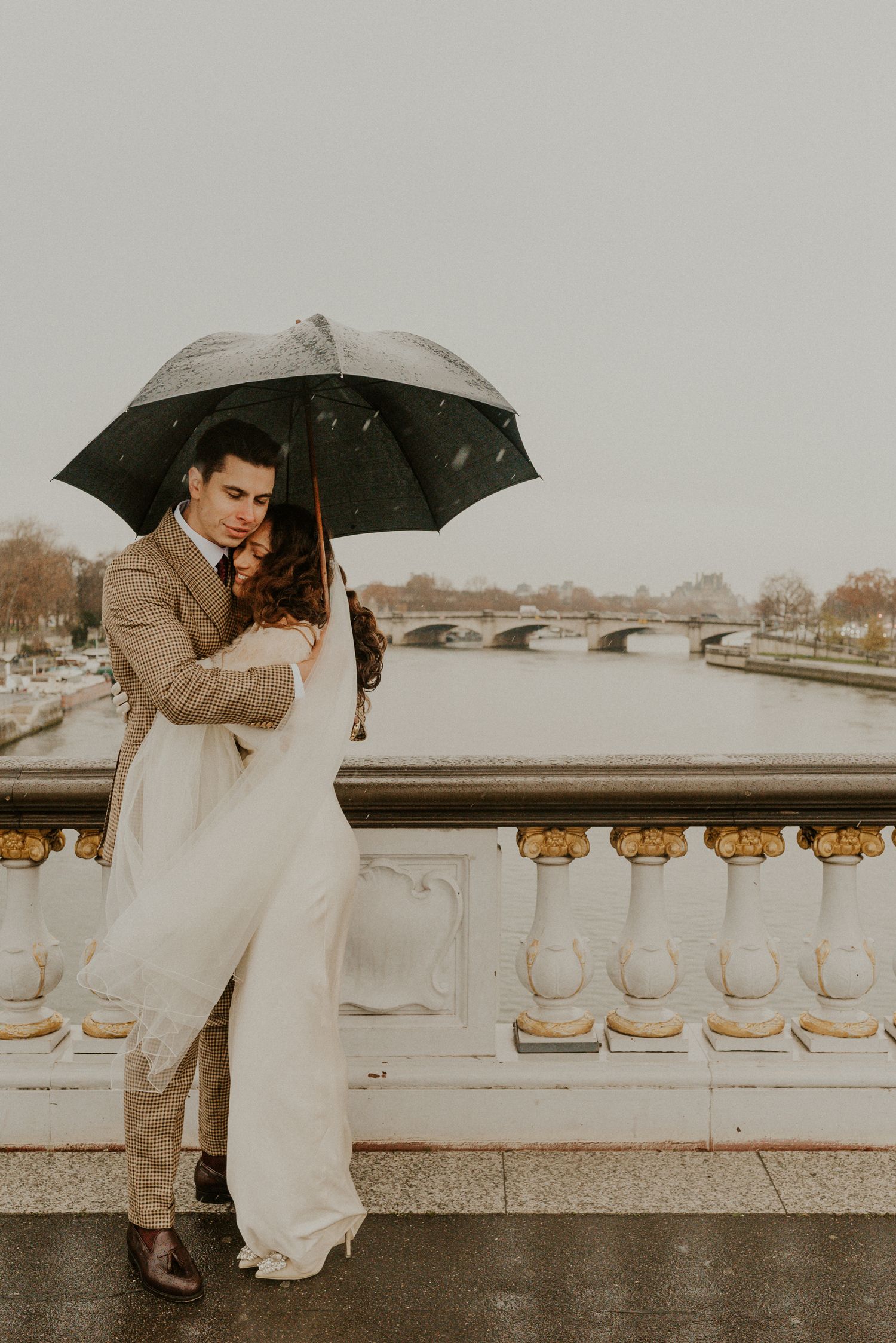 Couple sharing romantic moment on Paris bridge under umbrella on rainy day.