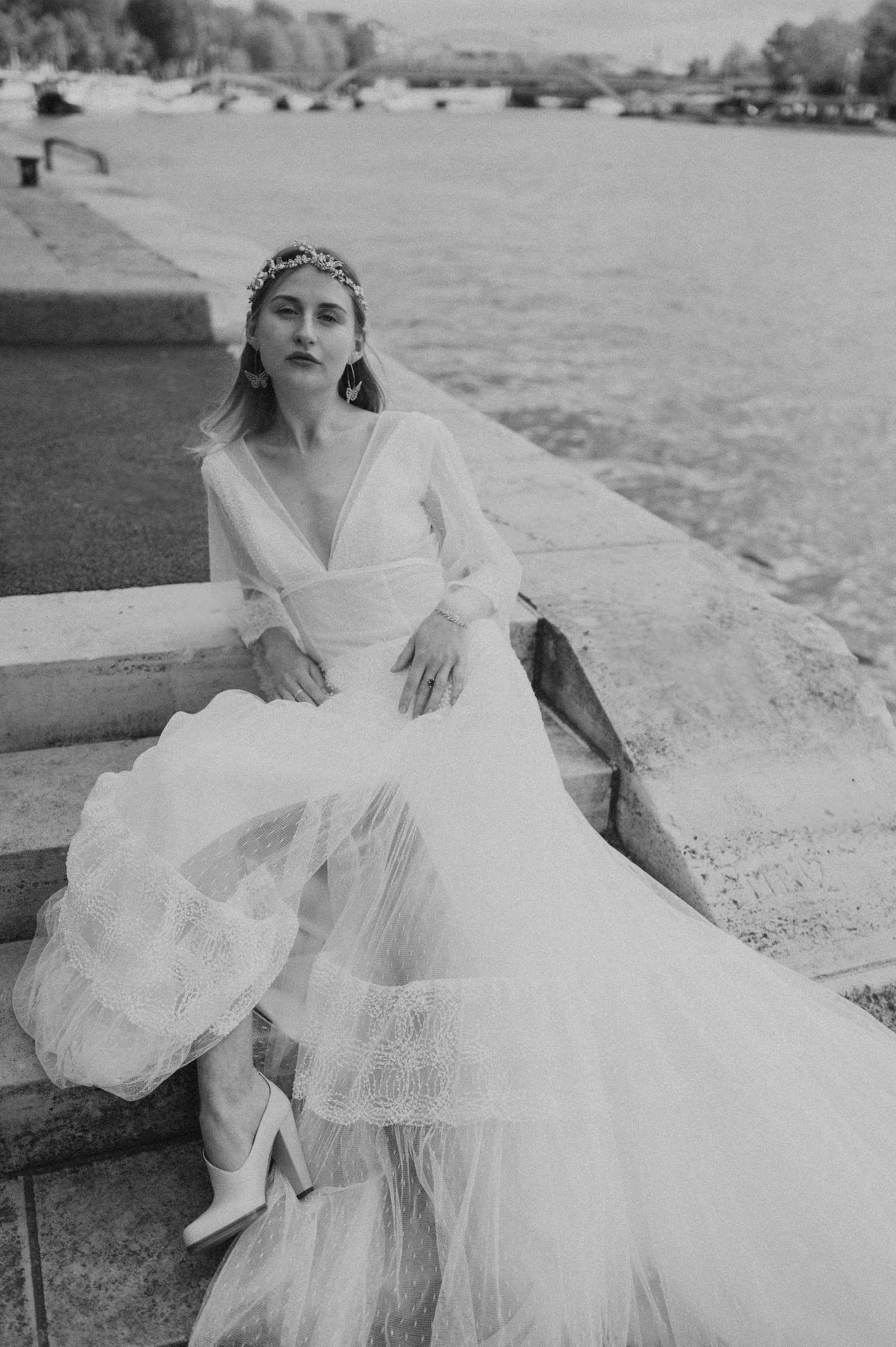 A black and white wedding photo of a bride lounging on stone steps by the water in a flowing tulle gown.