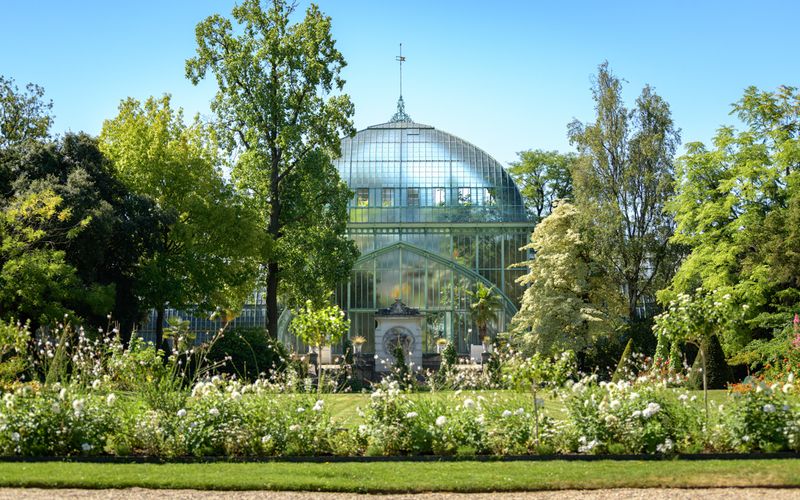Victorian-style glass greenhouse surrounded by lush summer gardens and tall trees.