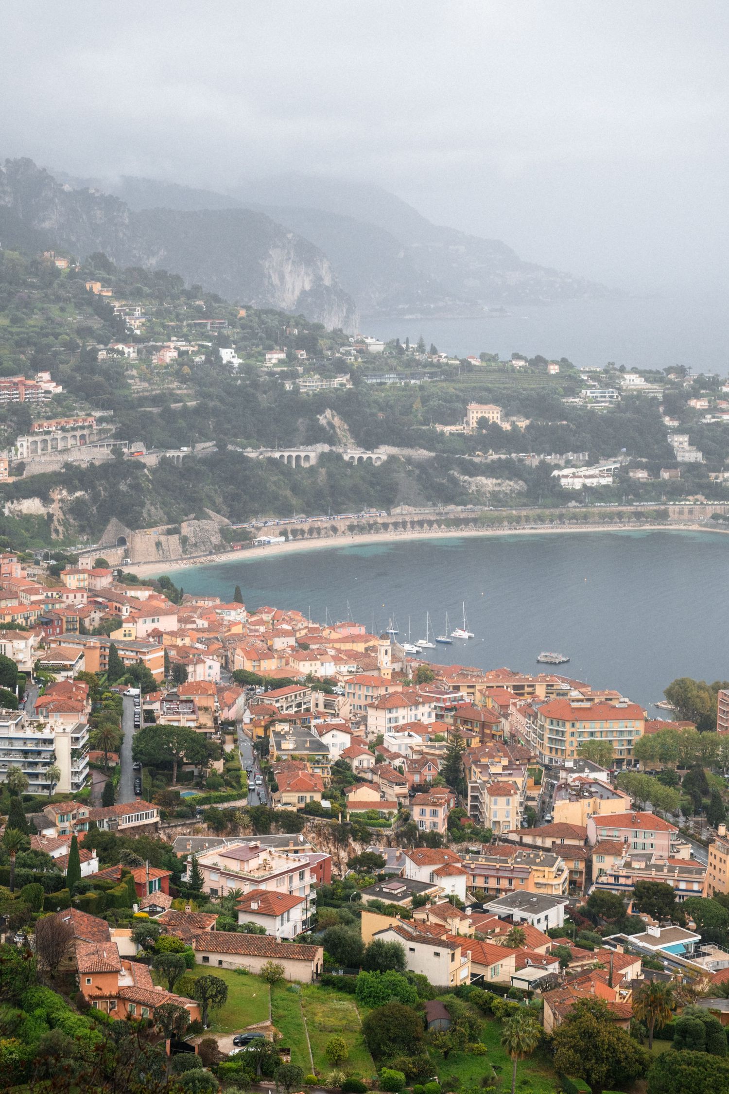 Aerial view of a Mediterranean coastal town with terracotta roofs nestled along a curved bay with mountains in the background.