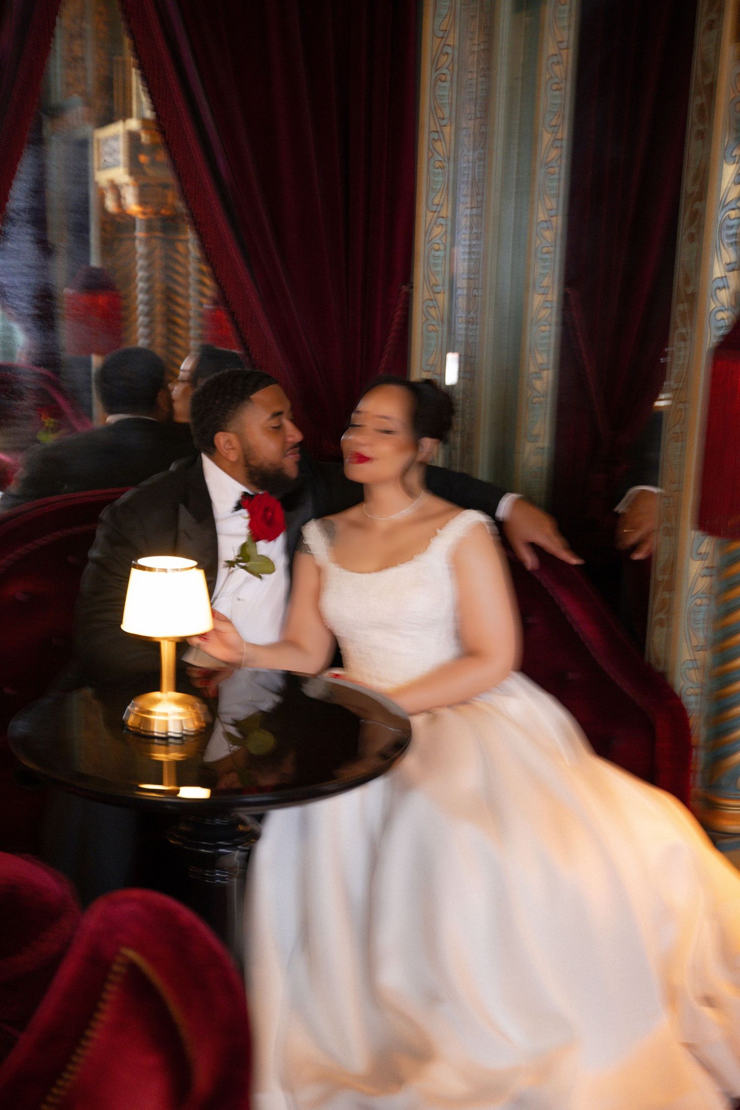 A bride and groom share a romantic moment seated together in an elegant venue with red velvet curtains and warm lamp light.