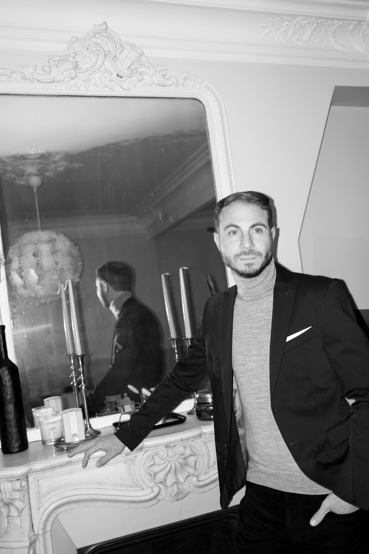 Black and white photo of a well-dressed man in blazer and turtleneck leaning against ornate fireplace mantel.