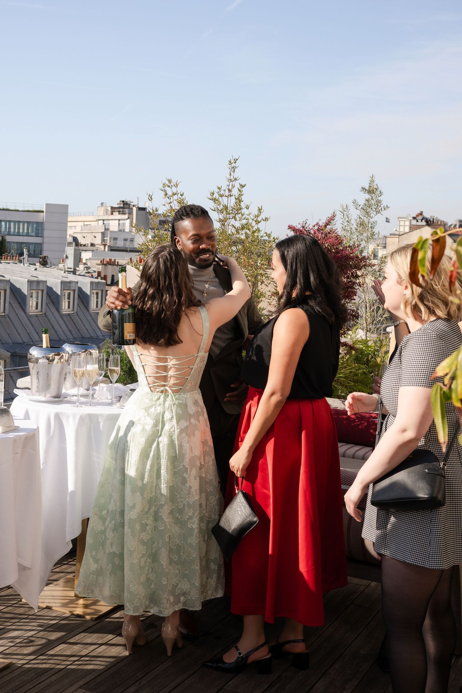 Elegant rooftop party with guests in colorful dresses socializing against a Parisian skyline on a sunny day.
