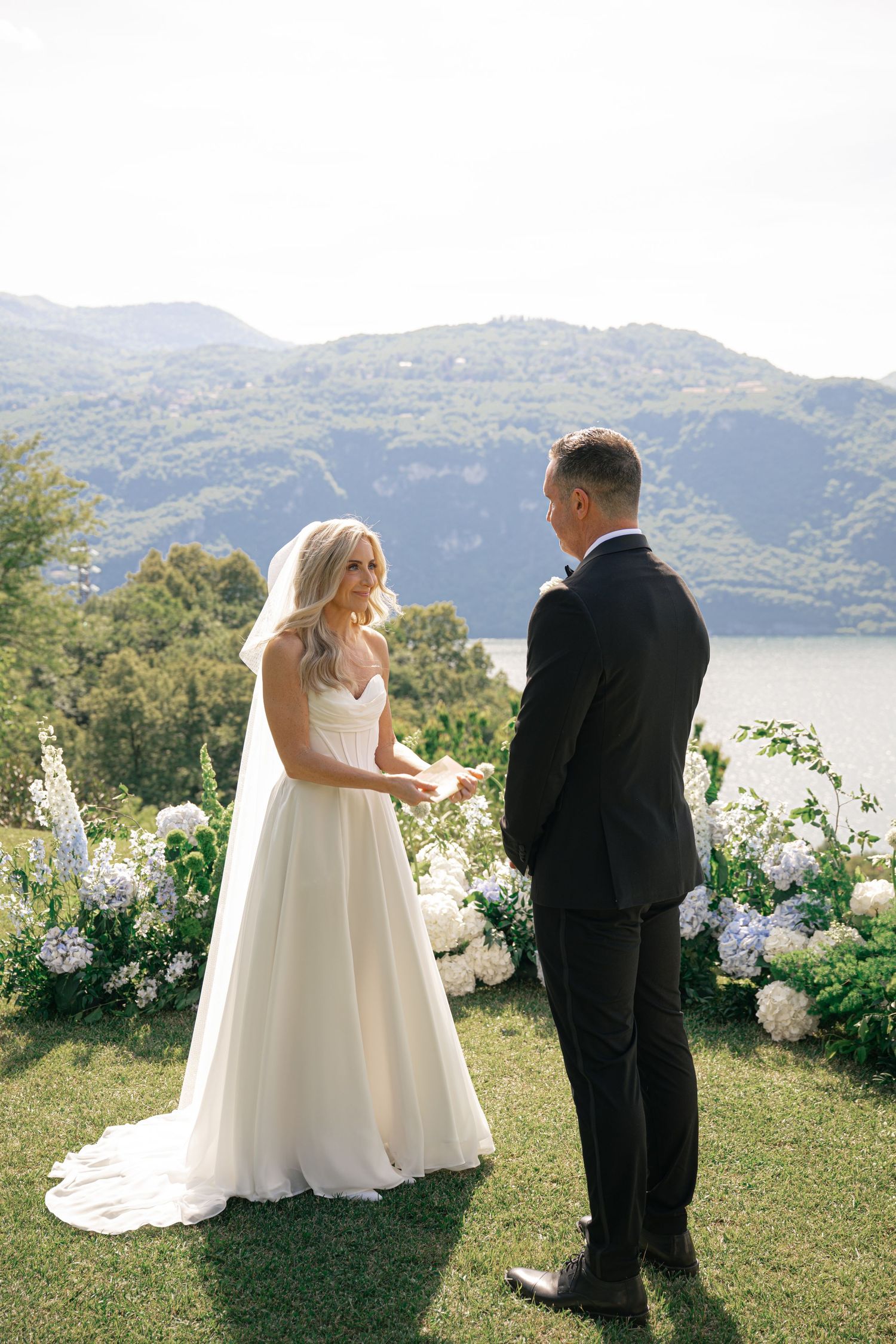 Bride and groom exchange vows outdoors with stunning mountain lake views and white hydrangea floral arrangements.