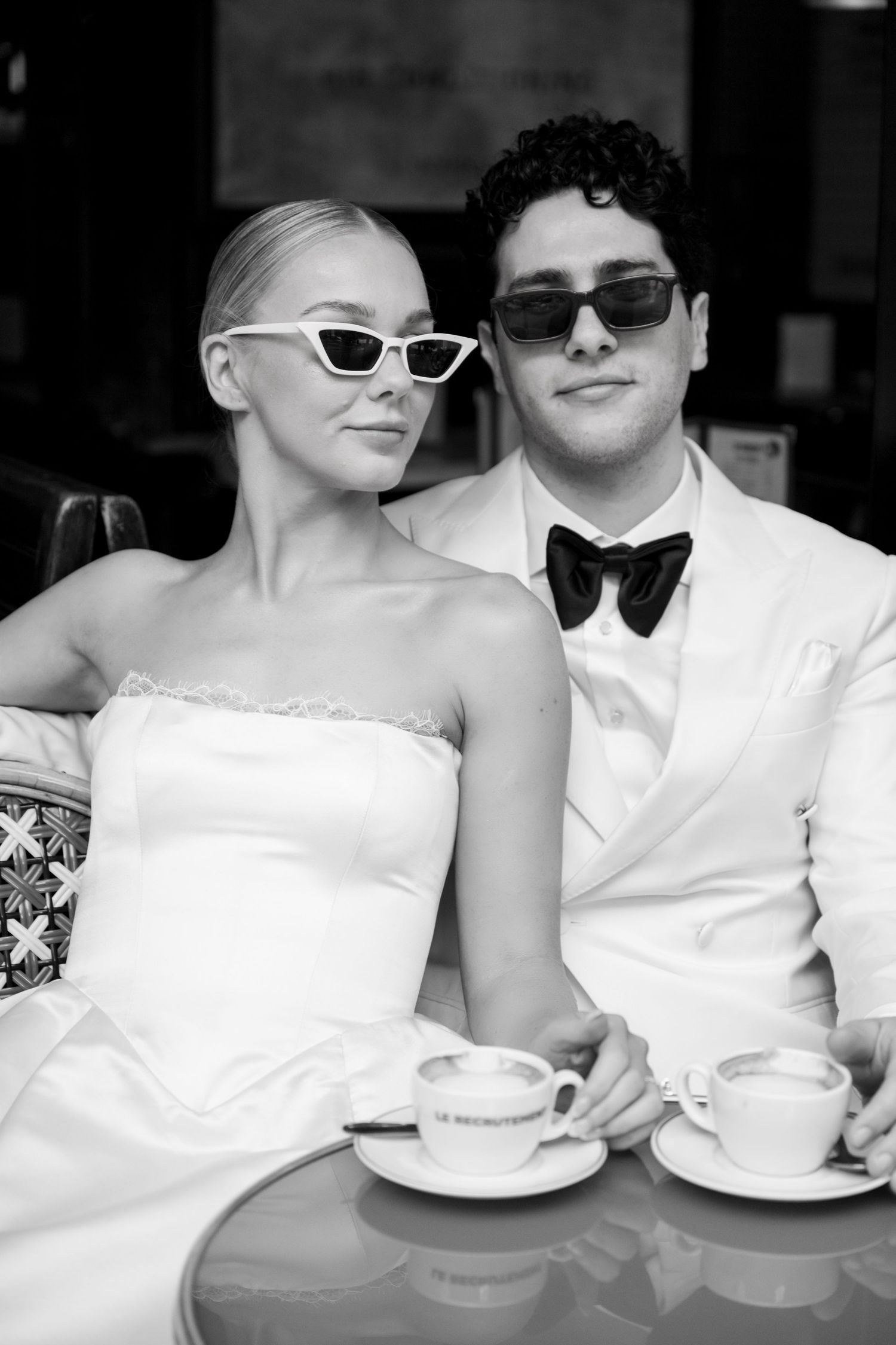 Stylish bride and groom in sunglasses sit at cafe table with espresso cups in black and white photo.