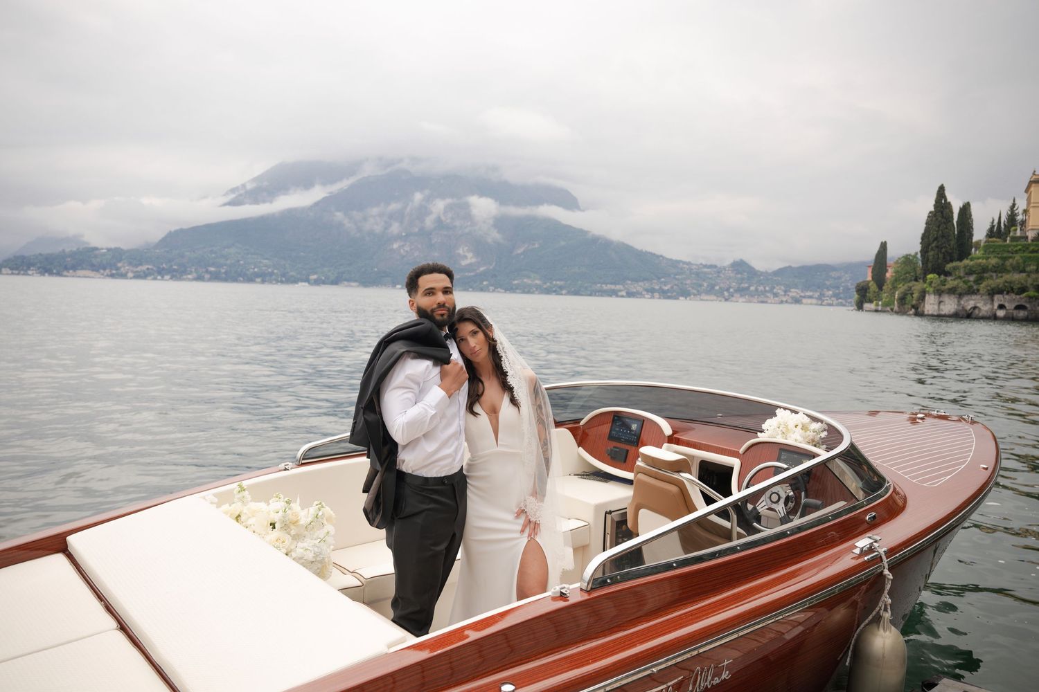 Newlyweds embrace on classic wooden speedboat on misty lake with mountain backdrop.
