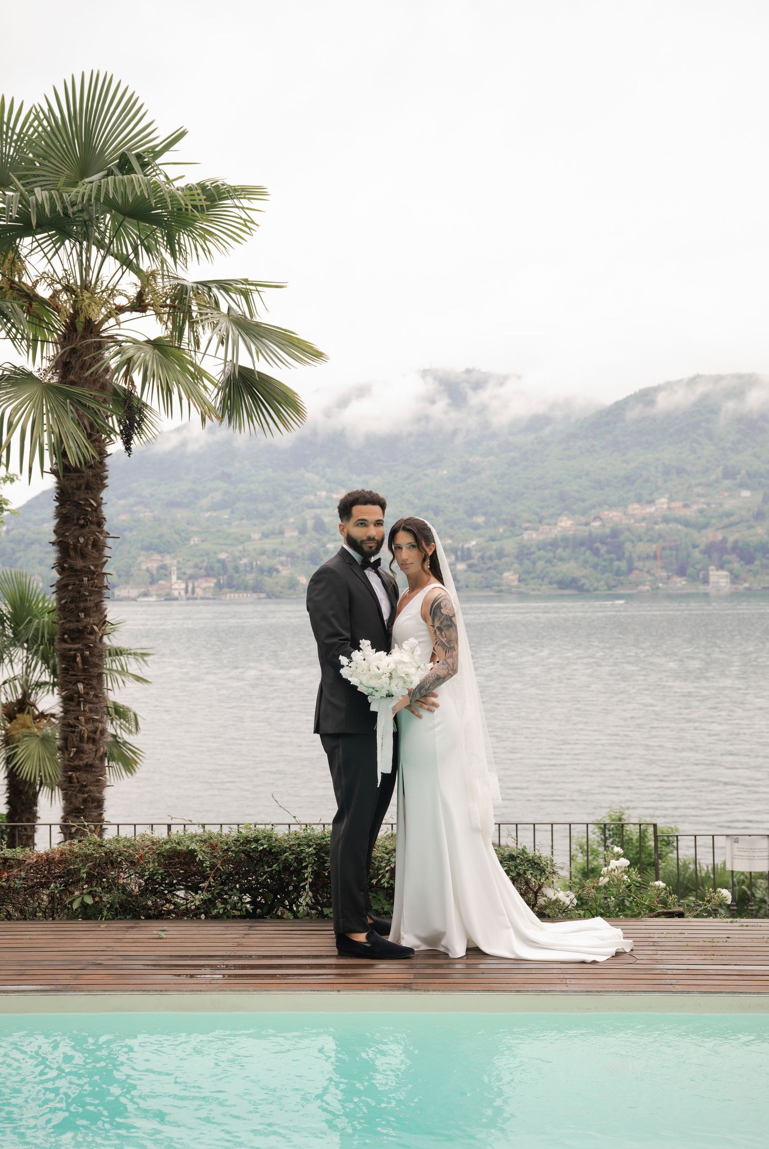 Bride and groom posing by a pool with Lake Como and palm trees in the misty background.