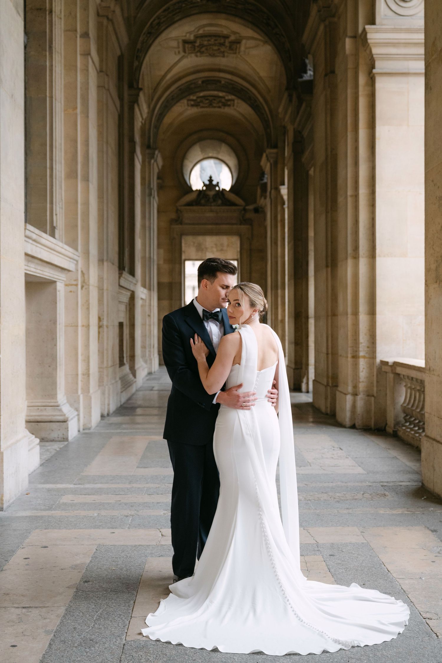A couple embraces in a grand colonnade with beautiful stone archways and natural lighting.