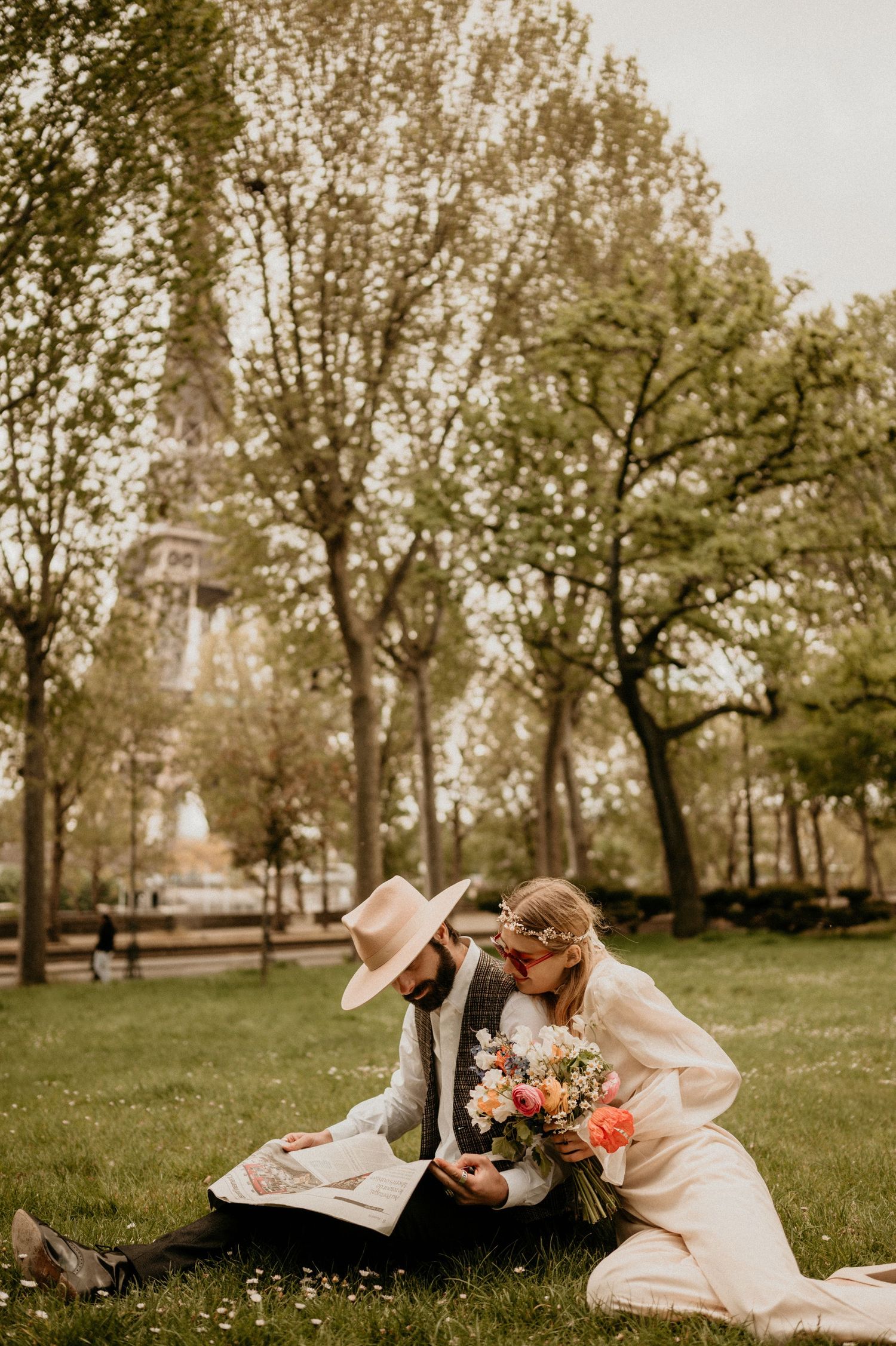 Intimate moment captured in park with couple sharing newspaper and flowers under spring trees.