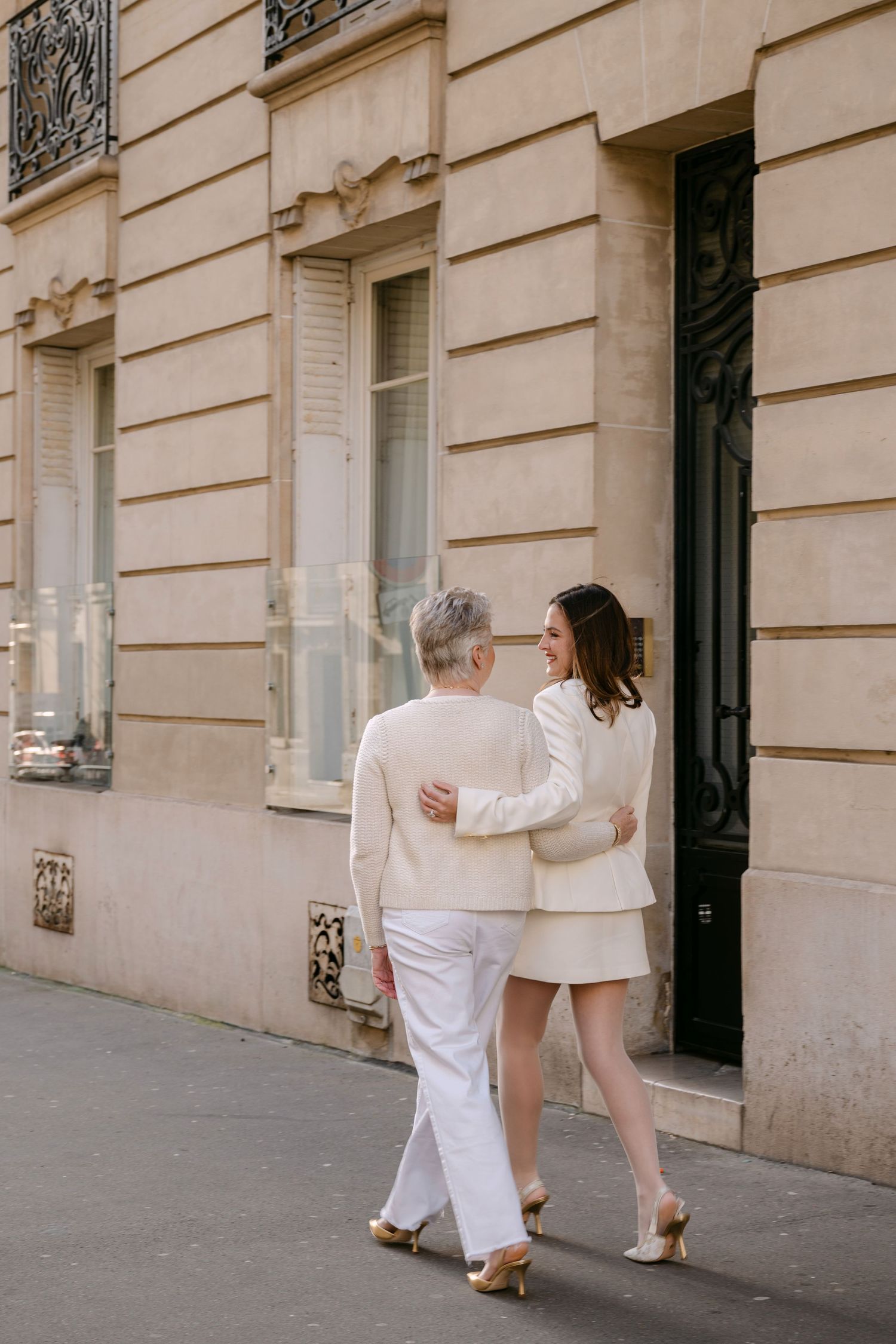 Two women in cream outfits walk arm in arm along classic Haussmann-style Parisian building facade.