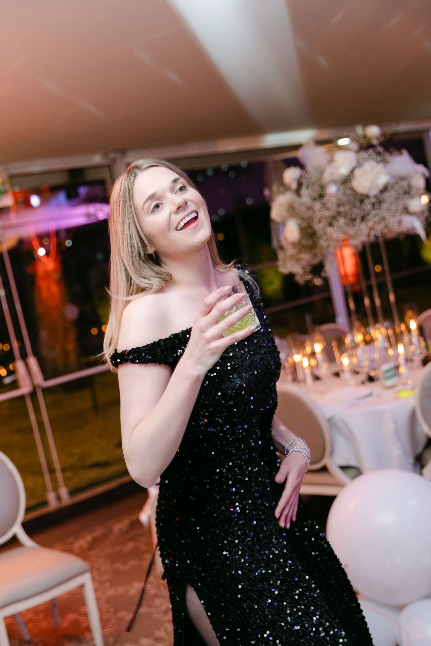 Elegant woman in black sequin off-shoulder gown holds cocktail at glamorous wedding reception.