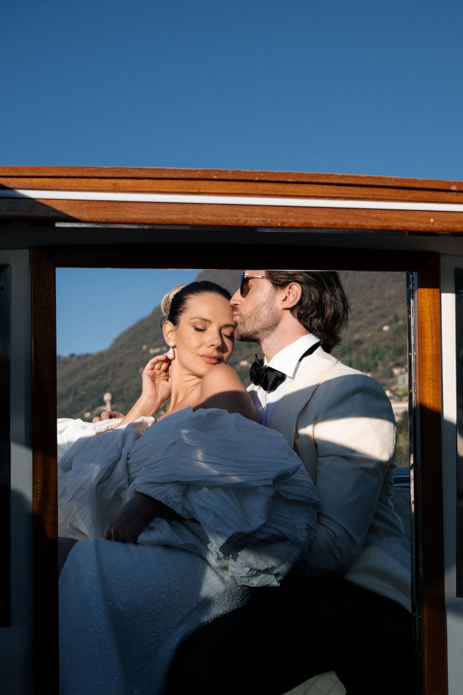 Romantic groom kissing bride through wooden boat window on scenic Italian lake.