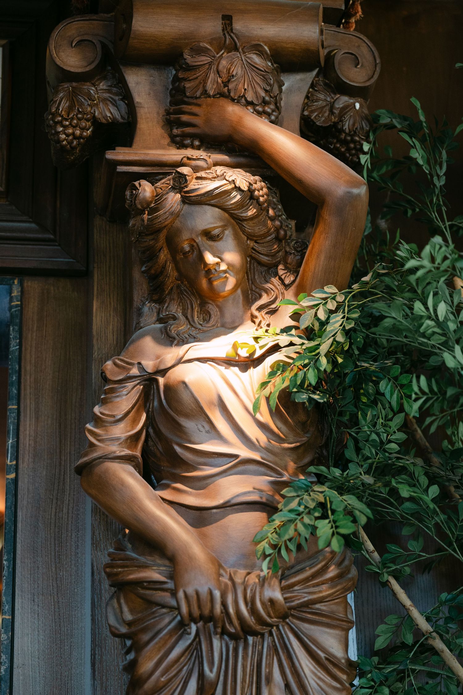 Ornate bronze sculpture of a classical female figure adorned with a floral crown beside lush green foliage.