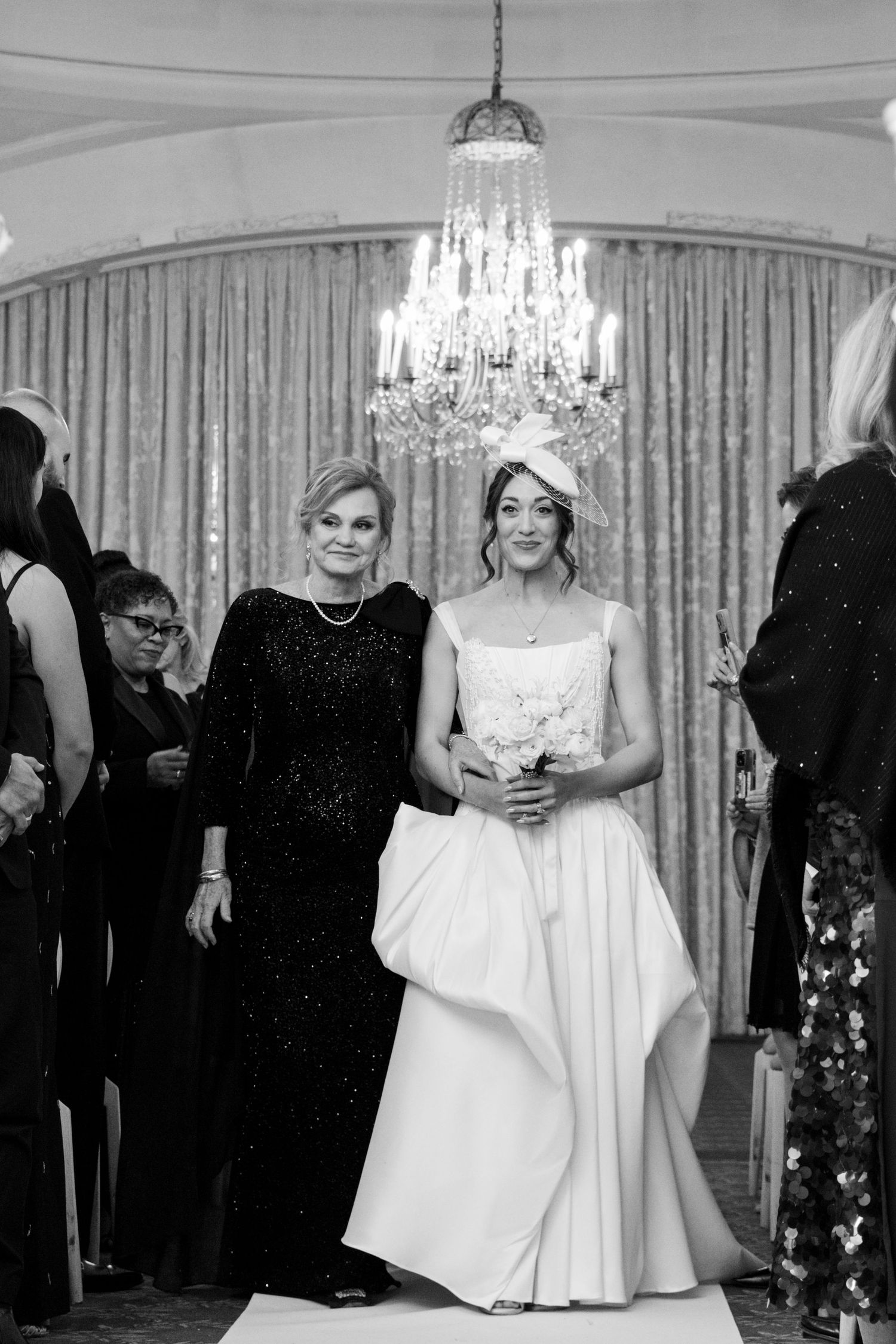 Black and white photo of bride in white gown with bouquet walking down aisle with mother in sequin dress.