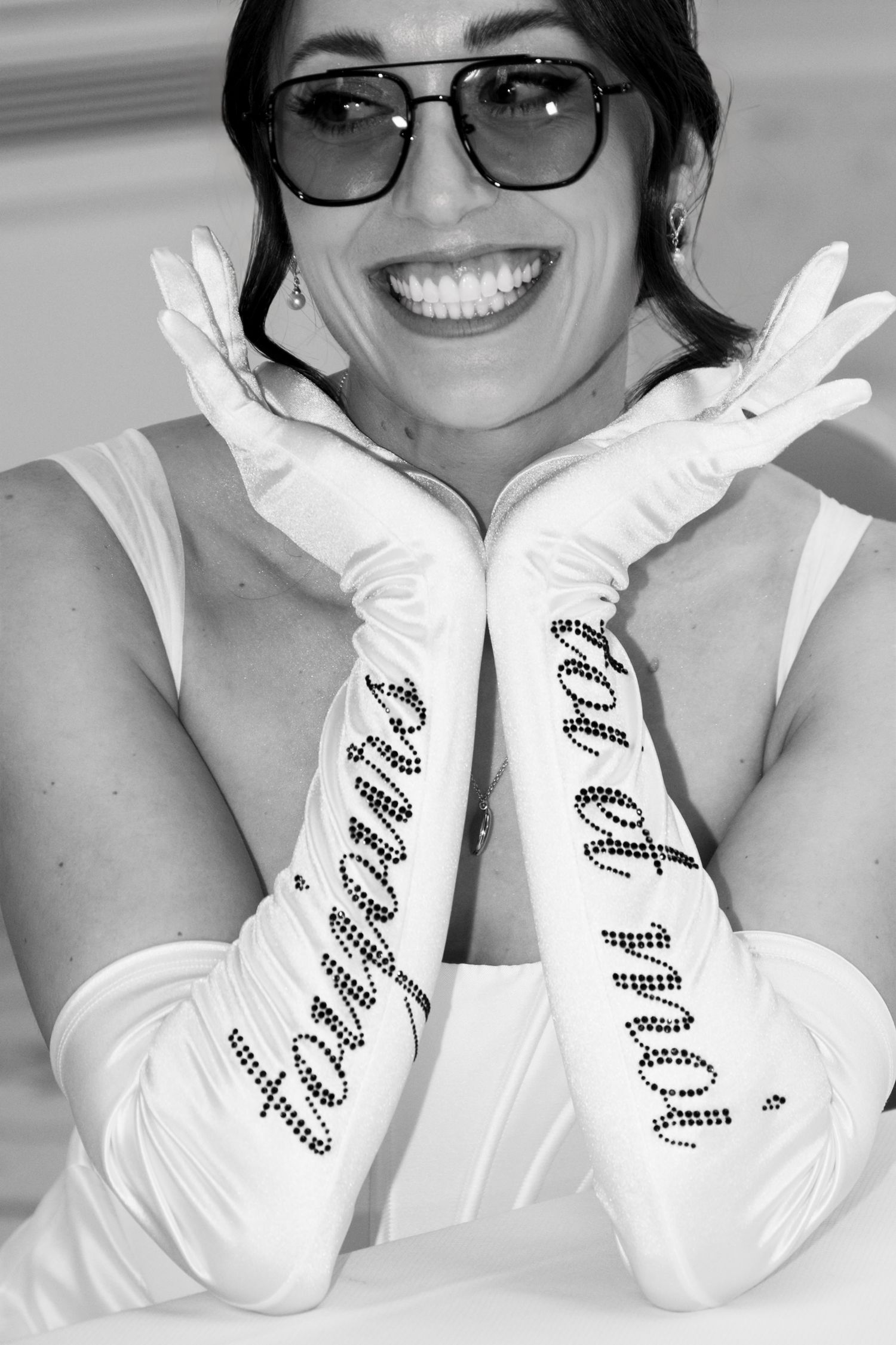 Smiling bride in white dress wearing long satin gloves with rhinestone script and oversized sunglasses.