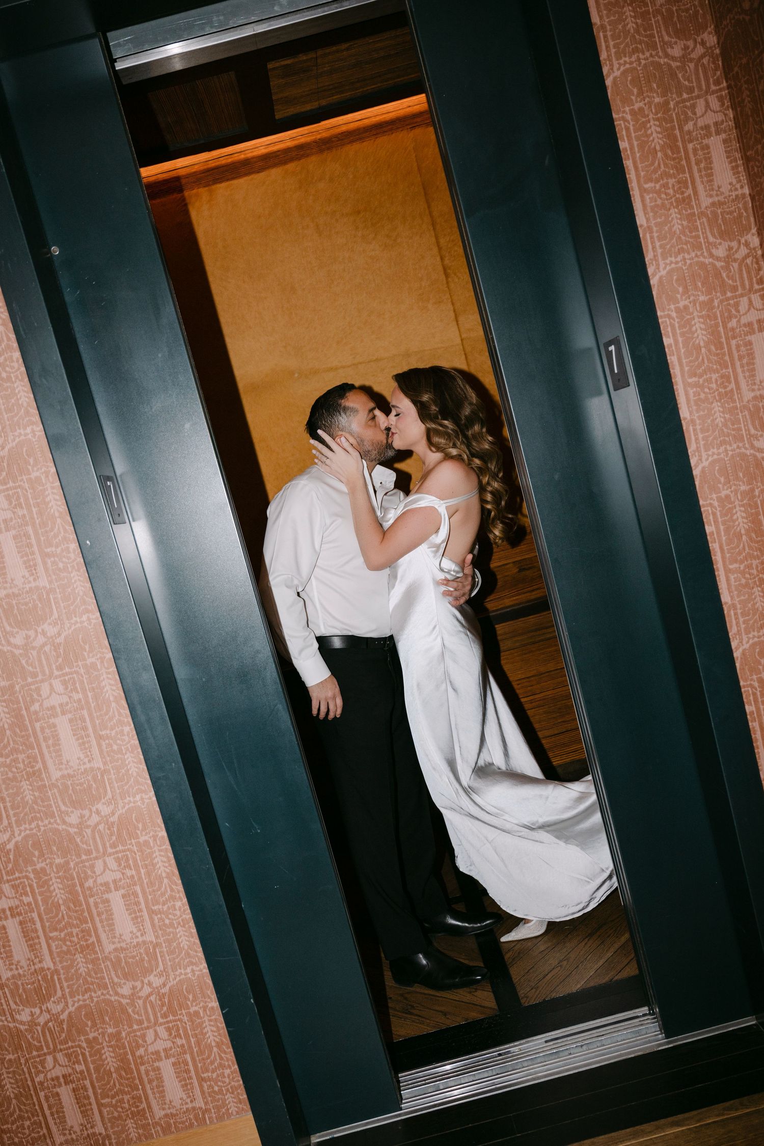 Romantic bride and groom sharing a kiss inside a golden hotel elevator in wedding attire.