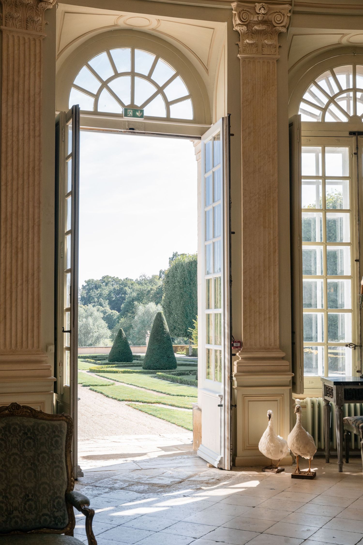 Grand French chateau interior with tall arched doors open to a formal garden with manicured topiary trees.