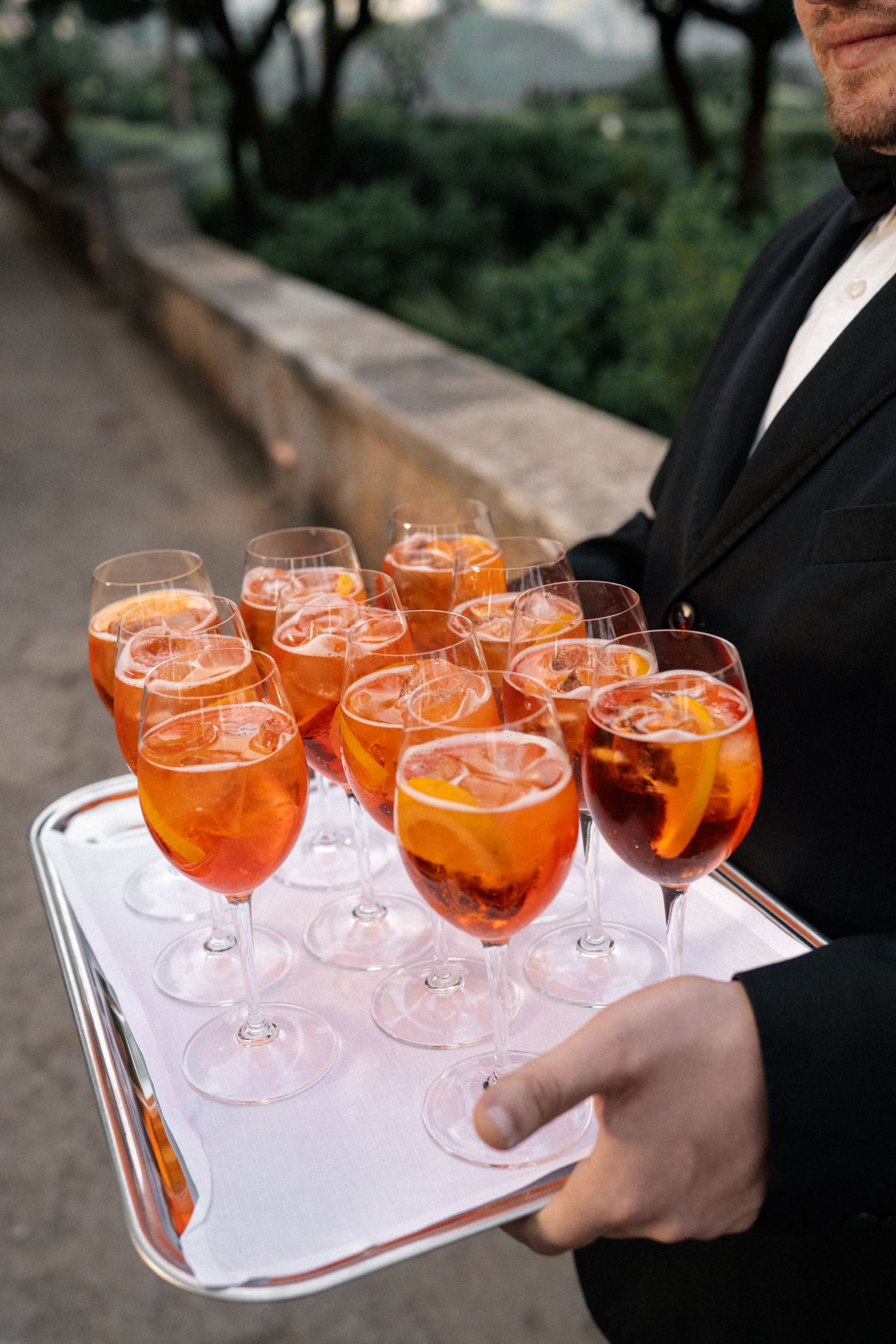 Waiter carries silver tray filled with Aperol Spritz cocktails garnished with orange slices at wedding.
