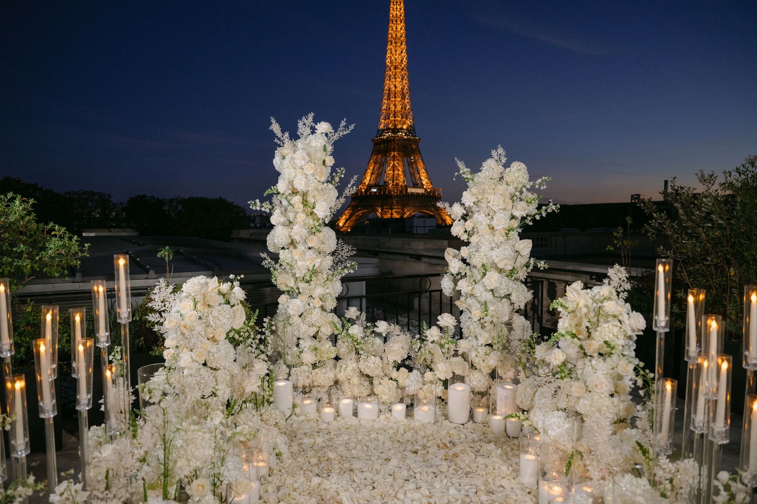 Romantic white floral arrangements and glowing candles set up for an evening proposal with the illuminated Eiffel Tower in the background.