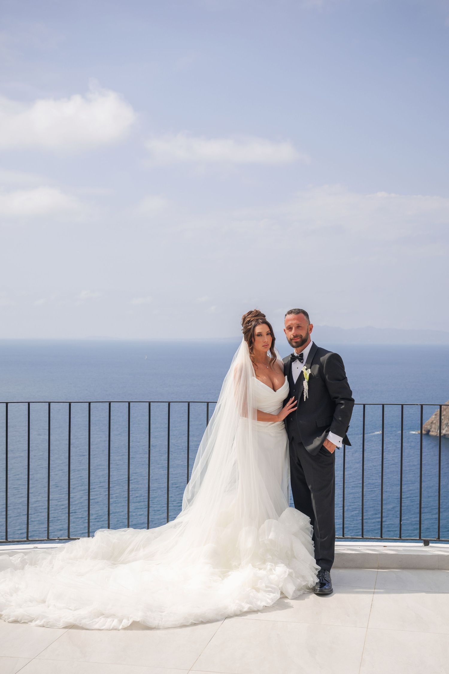 Bride and groom pose on seaside terrace, she in flowing white gown with veil, he in dark suit, ocean backdrop.