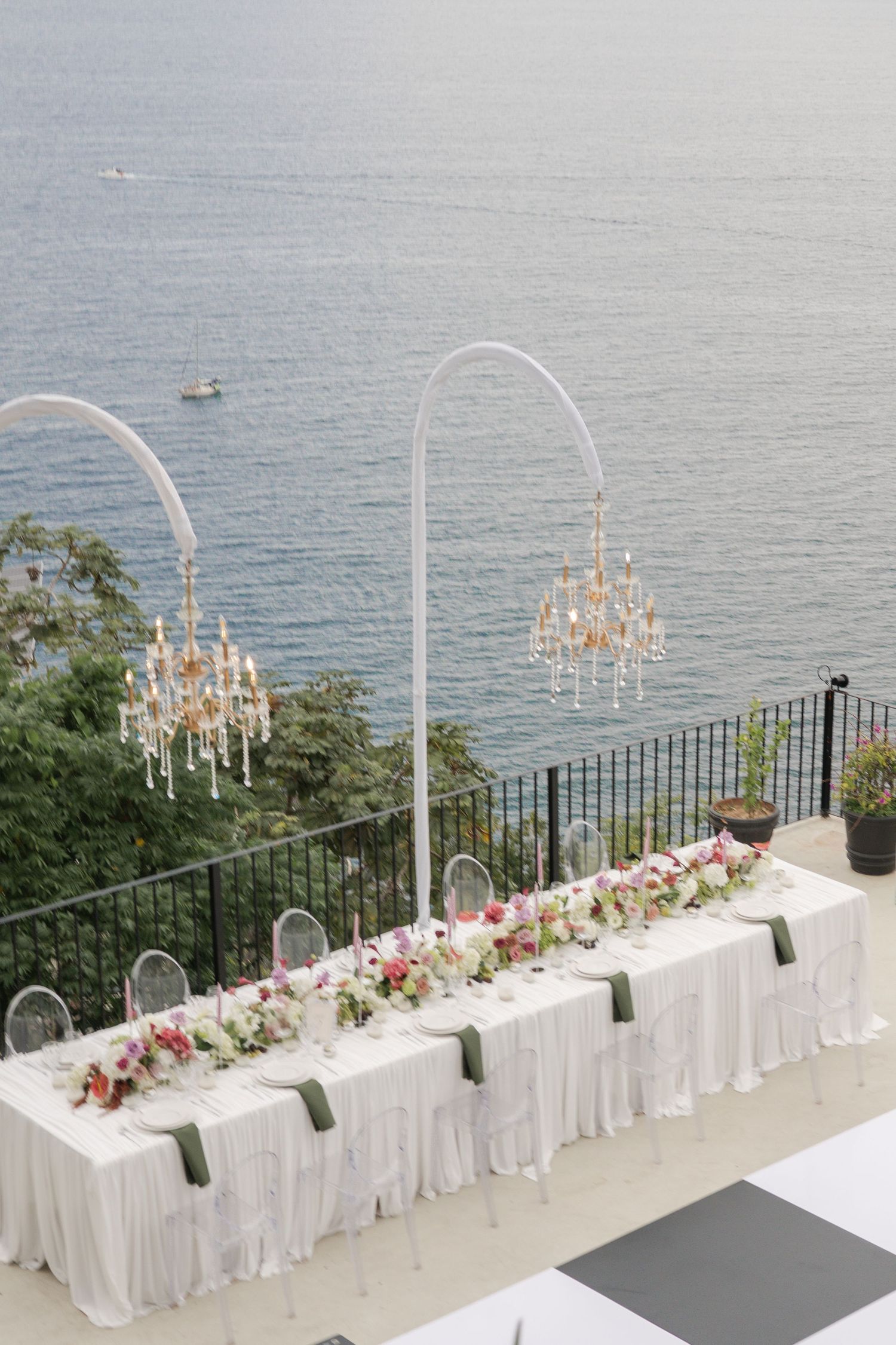 Elegant outdoor wedding reception table with chandeliers, white linens, pink floral centerpieces, and stunning ocean views.