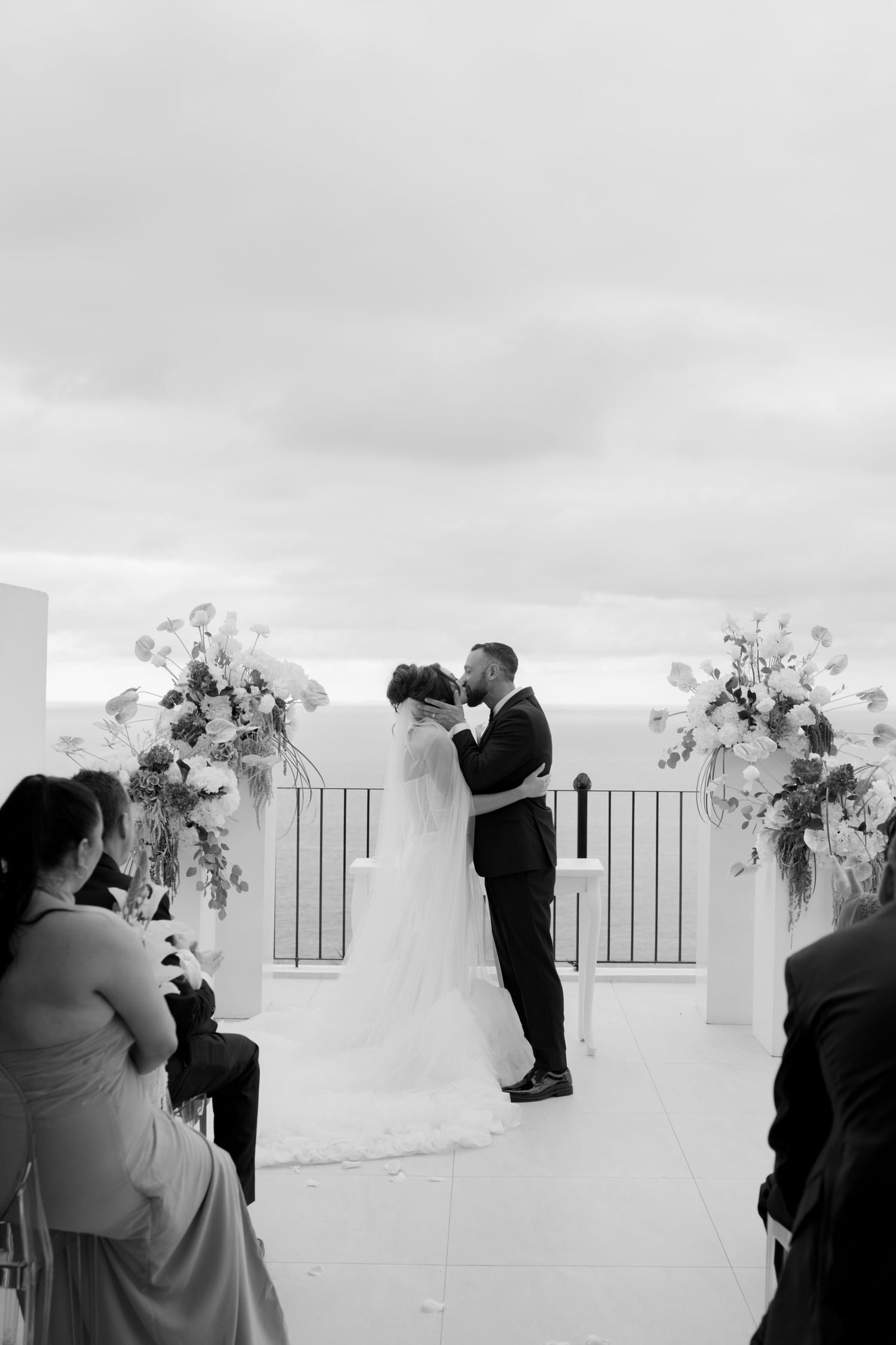 Black and white photo of a bride and groom sharing their first kiss at an outdoor wedding ceremony with floral arch.
