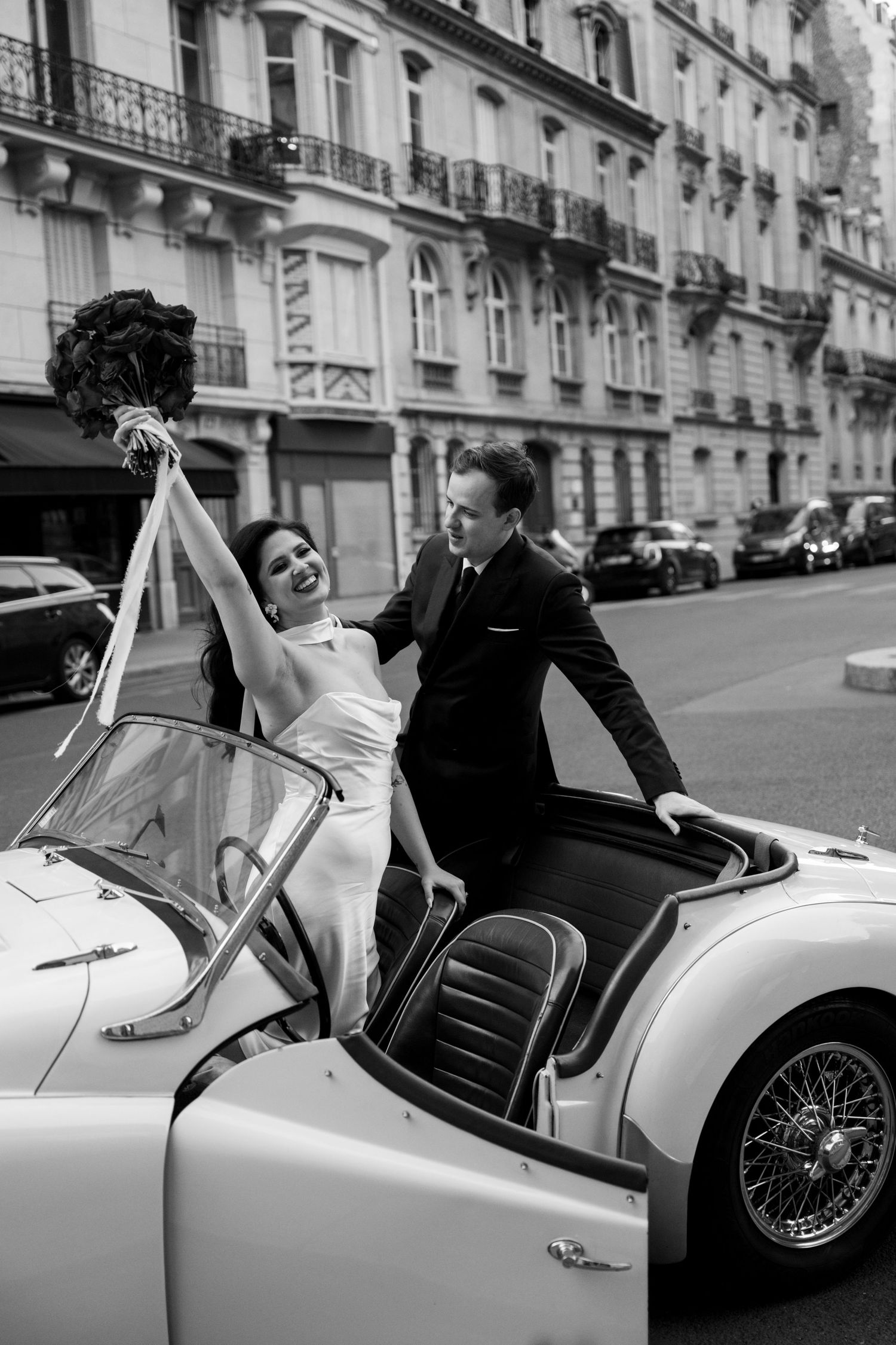 Bride and groom celebrating in a vintage white convertible on a Parisian street, black and white wedding photo.