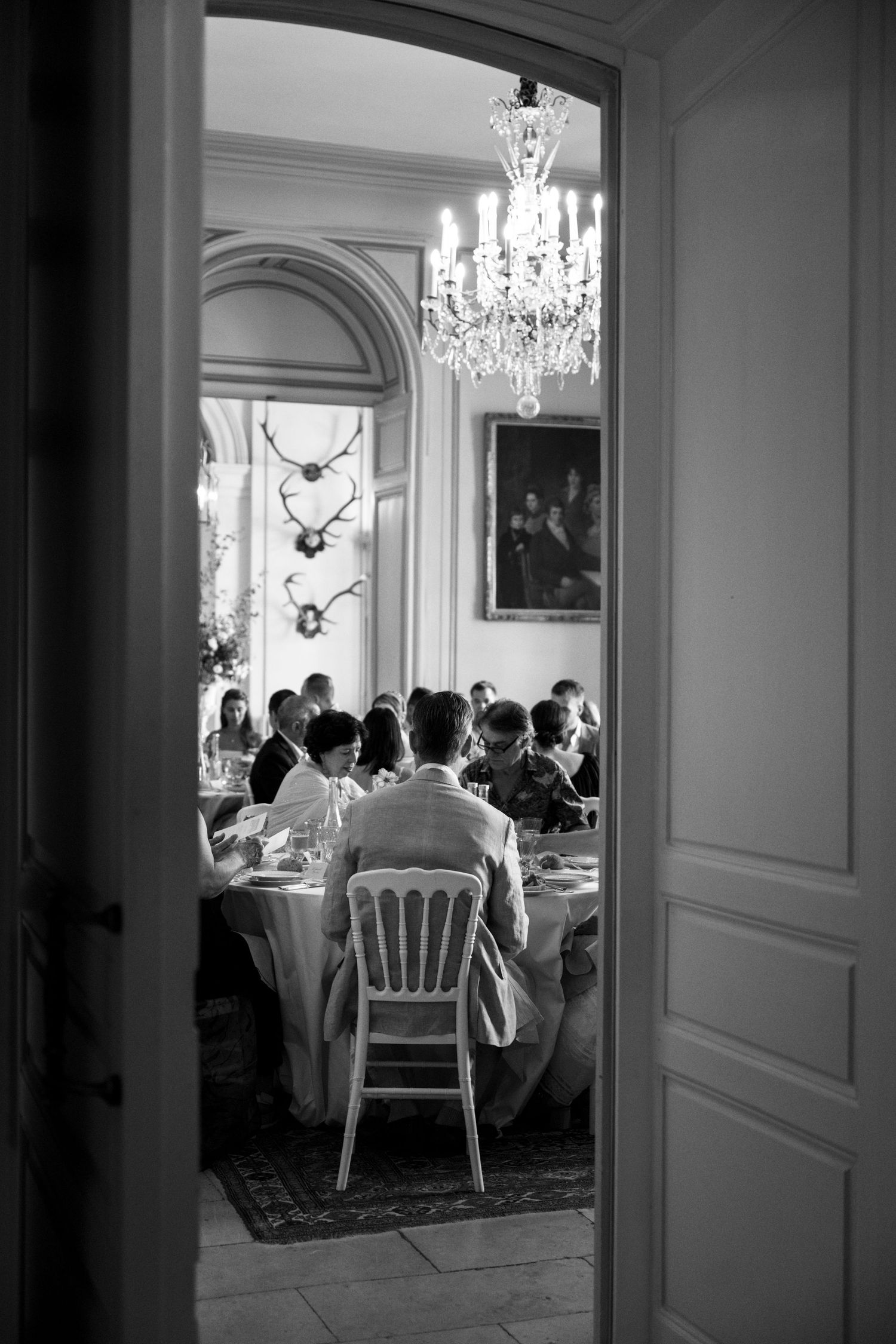 Black and white photo of wedding guests dining in a grand chateau room with crystal chandelier and antler wall decor.