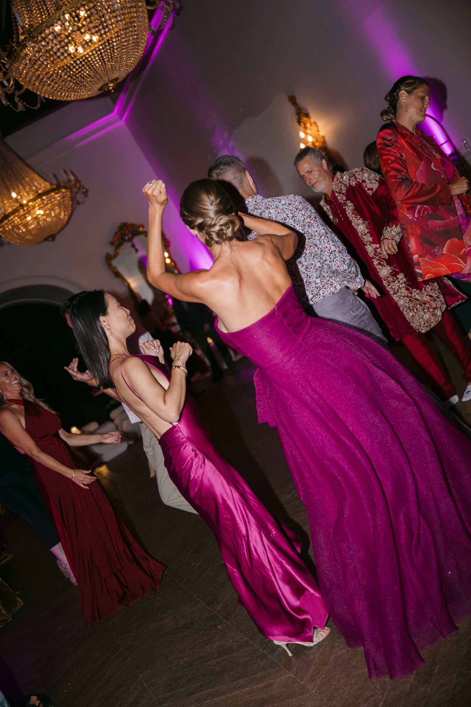 Wedding guests dancing energetically on a dance floor under golden chandeliers with purple lighting.