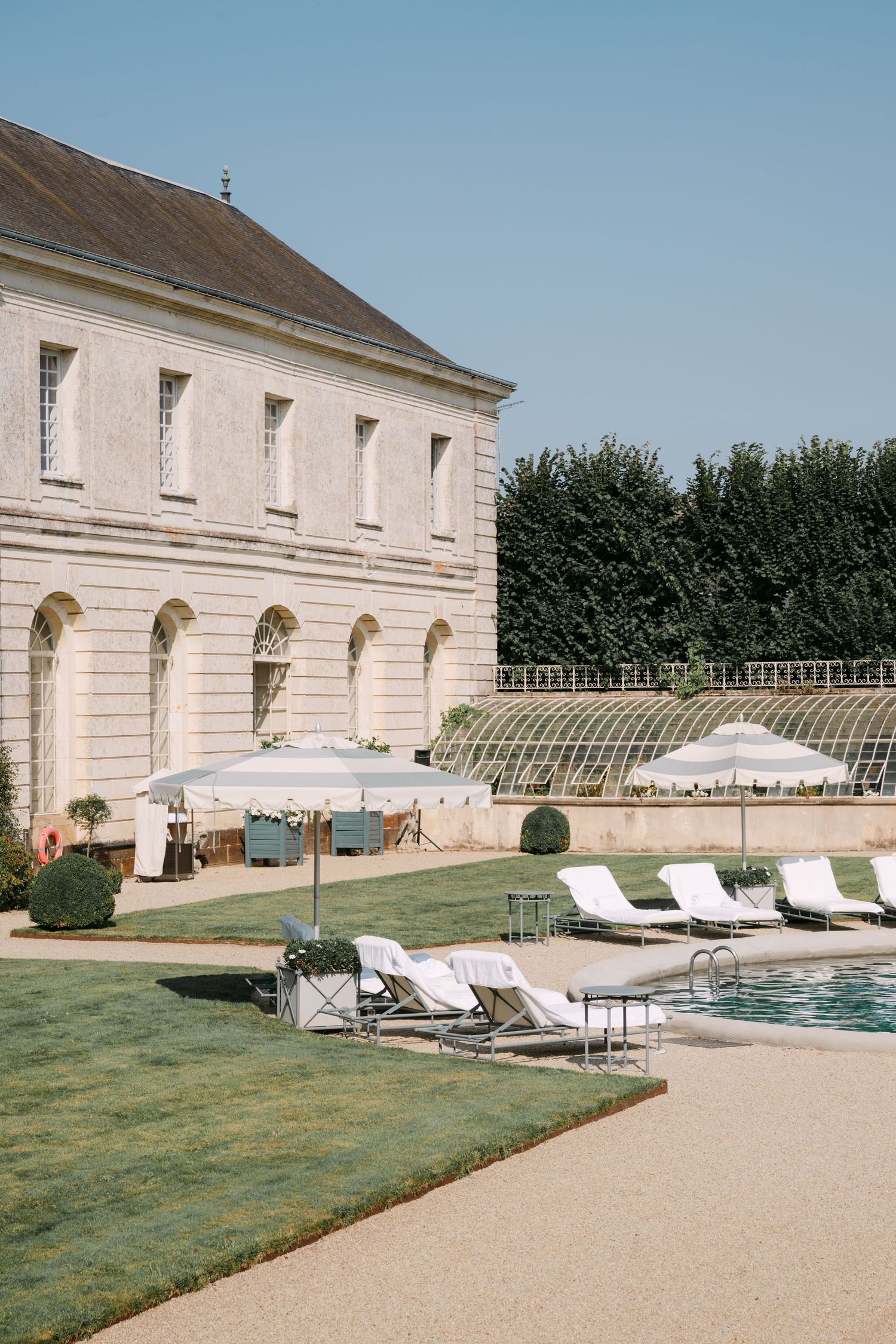 Classic French château with arched windows beside a pool with white loungers and greenhouse backdrop.