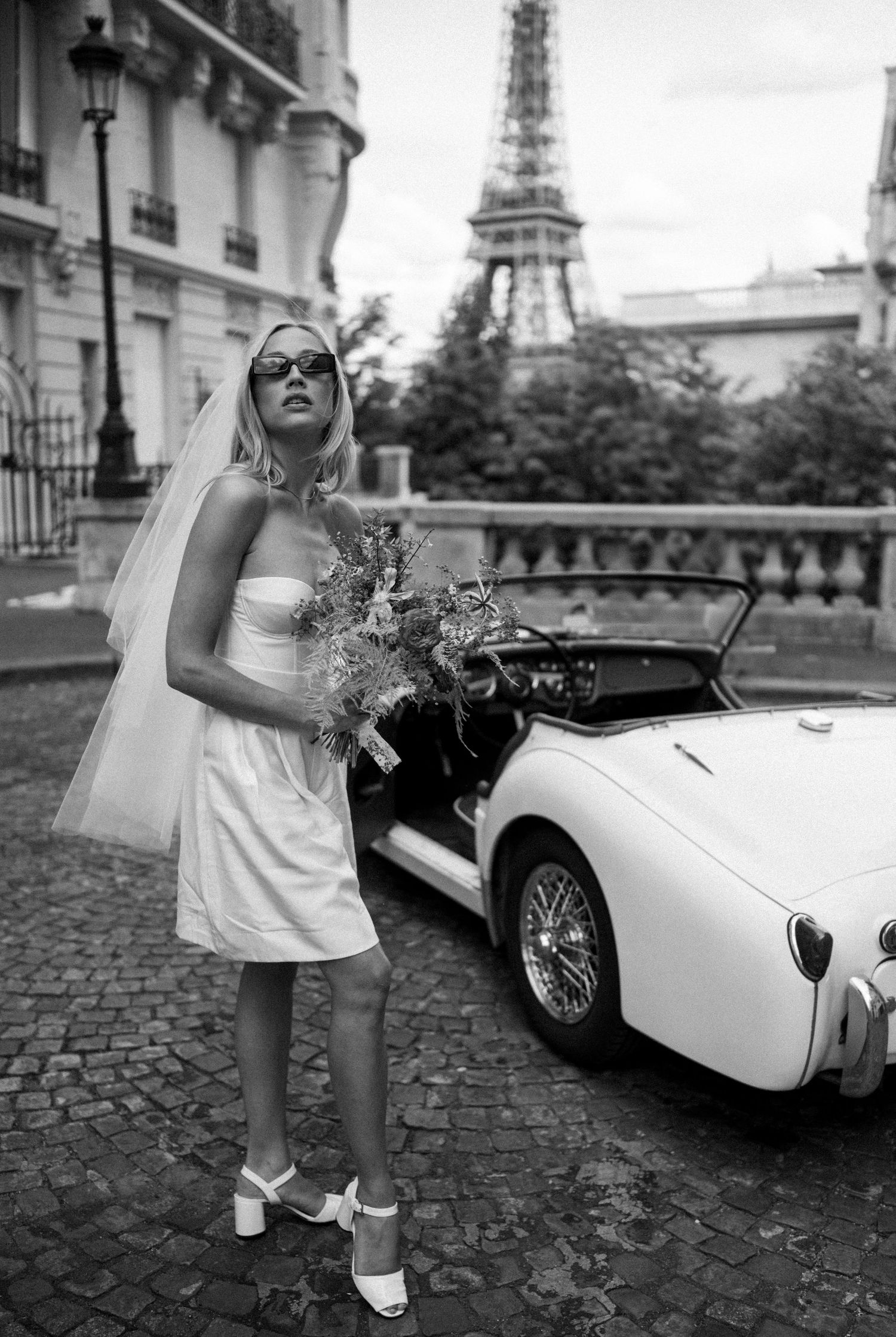Black and white photo of stylish bride in mini dress with sunglasses beside vintage Triumph car near Eiffel Tower.