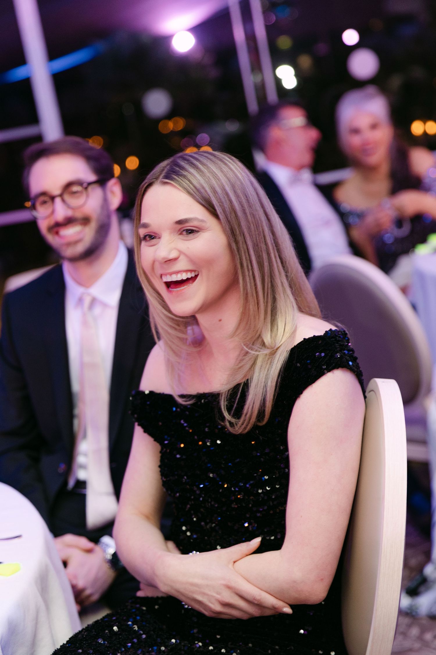 Laughing blonde woman in a black sequin dress enjoying a wedding reception dinner in a decorated venue.